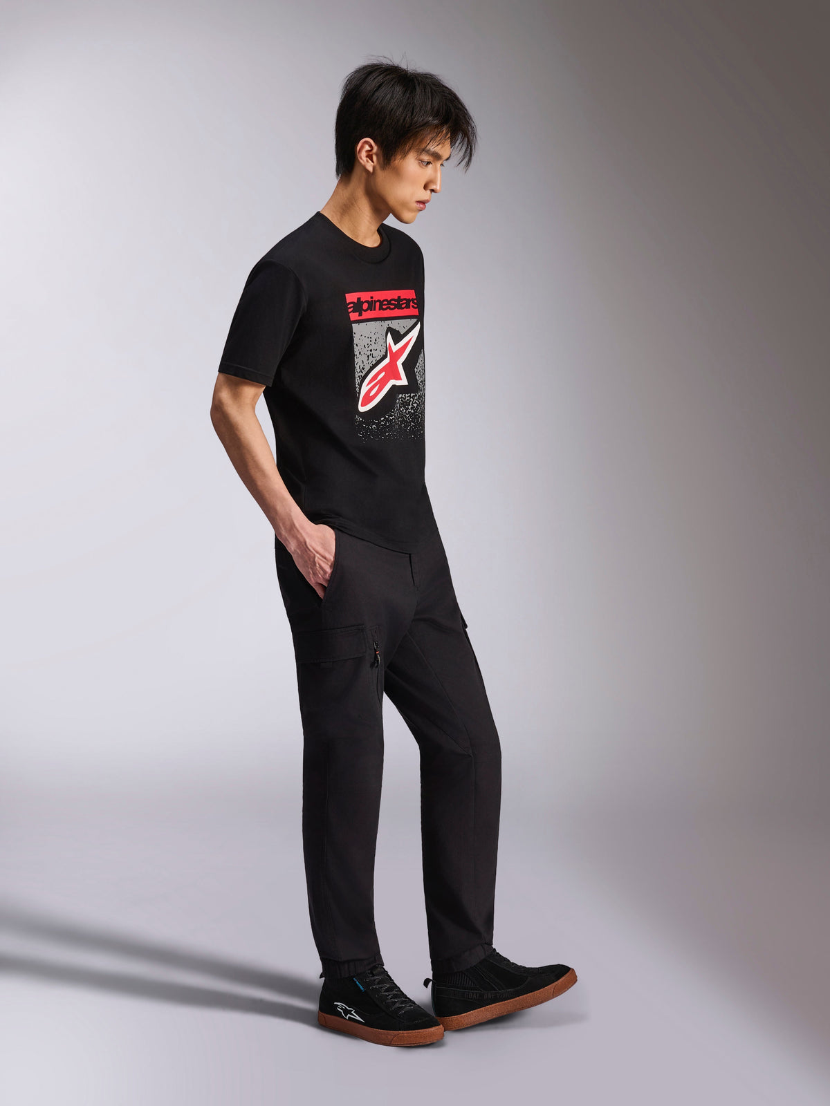 Alpinestars Noka CSF Tee, kurzärmeliges T-Shirt, Schwarz, Front Astars Logo mit rot-weißem MX-Verlauf, getragen mit schwarzer Cargohose und Sneakern, lässige, vom Motorsport inspirierte Streetwear