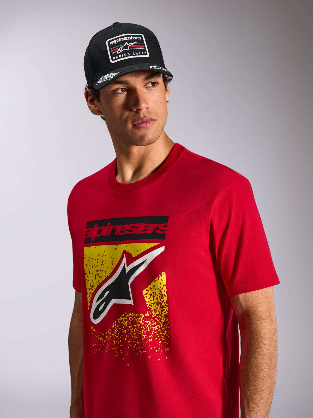 Alpinestars Noka CSF Tee, T-shirt met korte mouwen, rood, met een Astars-logo op de voorzijde met geel en rood MX-verloop, gedragen met een zwarte Alpinestars 'Racing Ahead' pet, casual motorsport streetwear