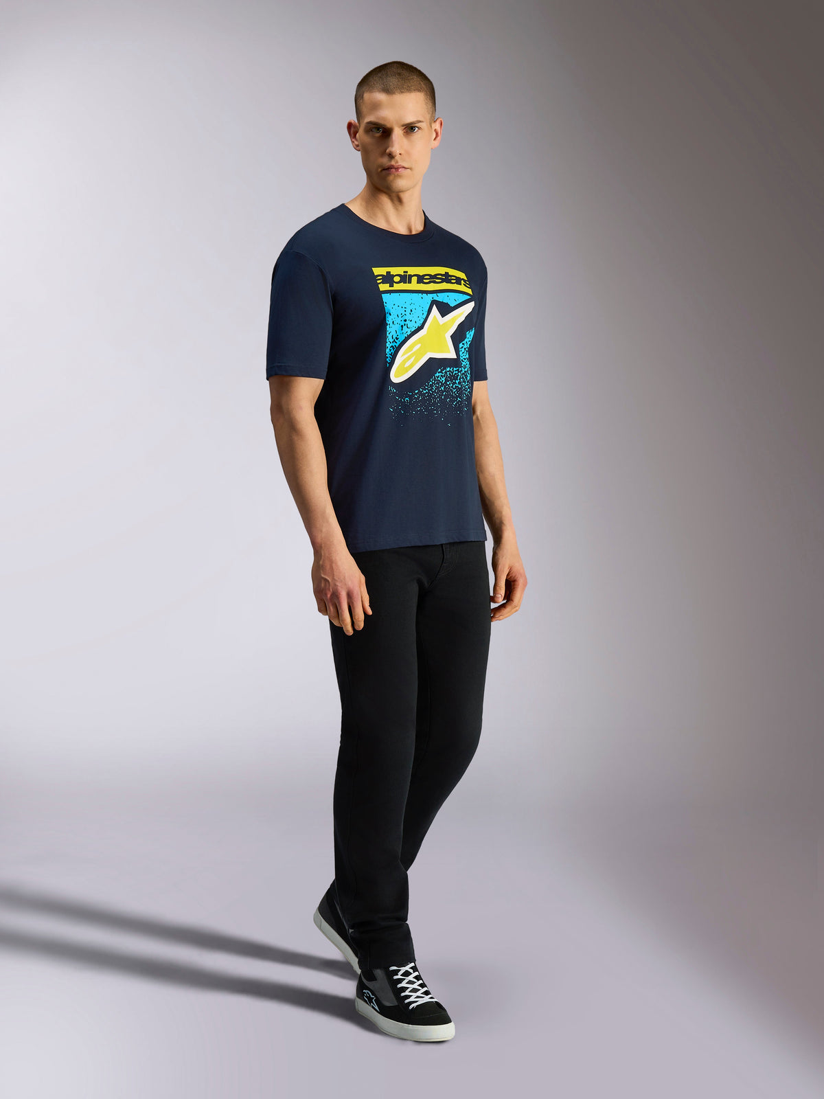 T-shirt Alpinestars Noka CSF, t-shirt à manches courtes, bleu marine, logo Astars jaune avec graphique en dégradé bleu MX, mannequin portant un t-shirt marine avec un pantalon noir et des baskets, streetwear décontracté motorsport