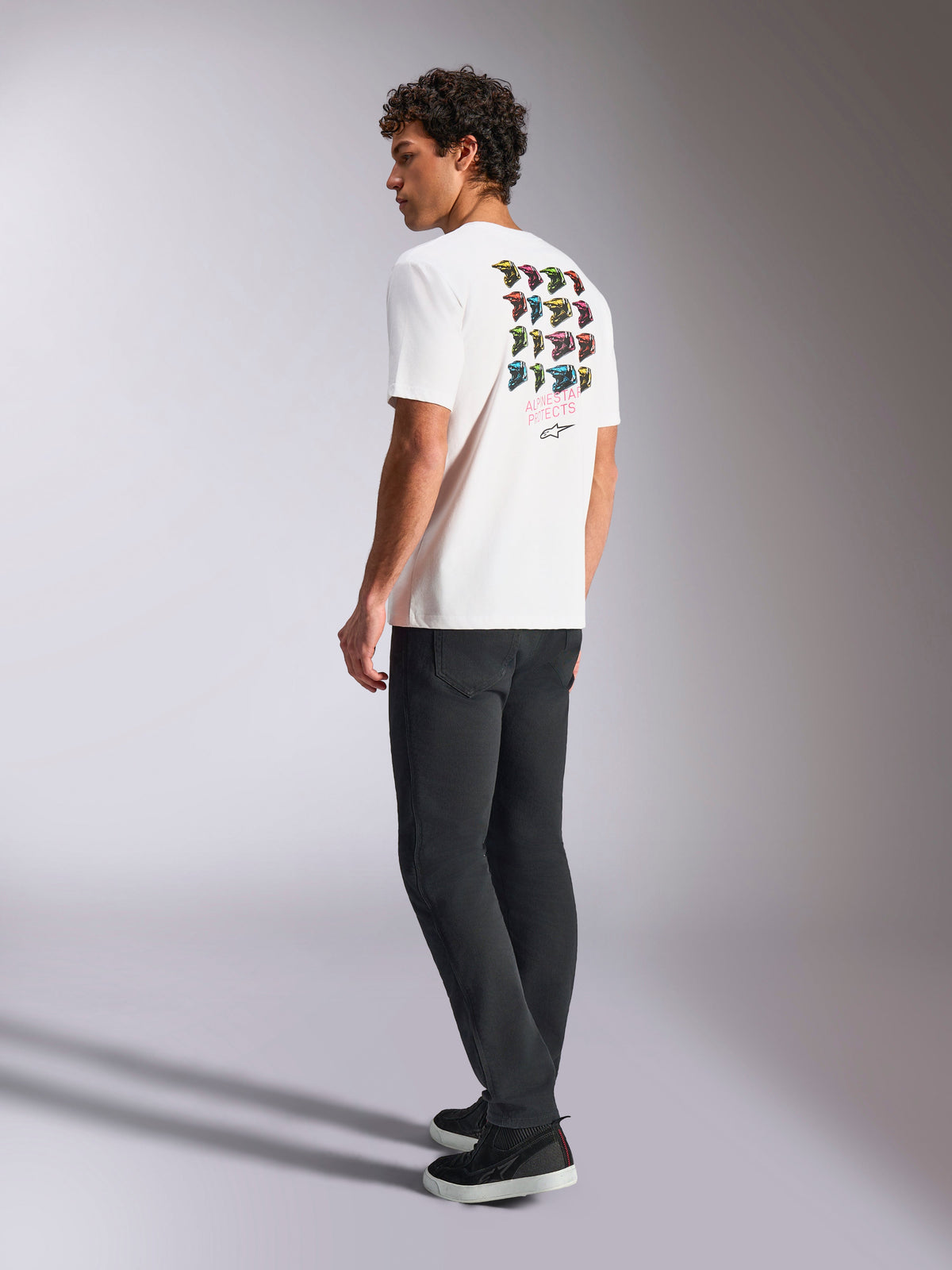 T-shirt Alpinestars Pop Helmet CSF, t-shirt à manches courtes, blanc, vue de dos sur un mannequin présentant un graphisme coloré du casque M10, porté avec un pantalon sombre et des baskets noires, streetwear décontracté inspiré des sports mécaniques