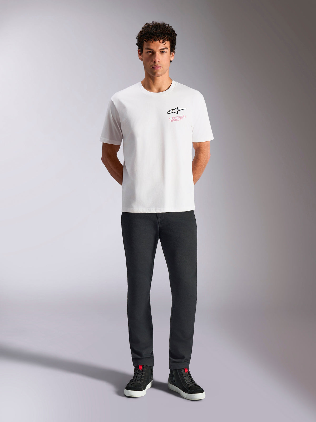 Alpinestars Pop Helmet CSF Tee, t-shirt à manches courtes, blanc, porté par un mannequin avec un pantalon noir et des baskets montantes noires, logo Astars à l'avant avec texte rose « Alpinestars Protects », streetwear motorsport décontracté