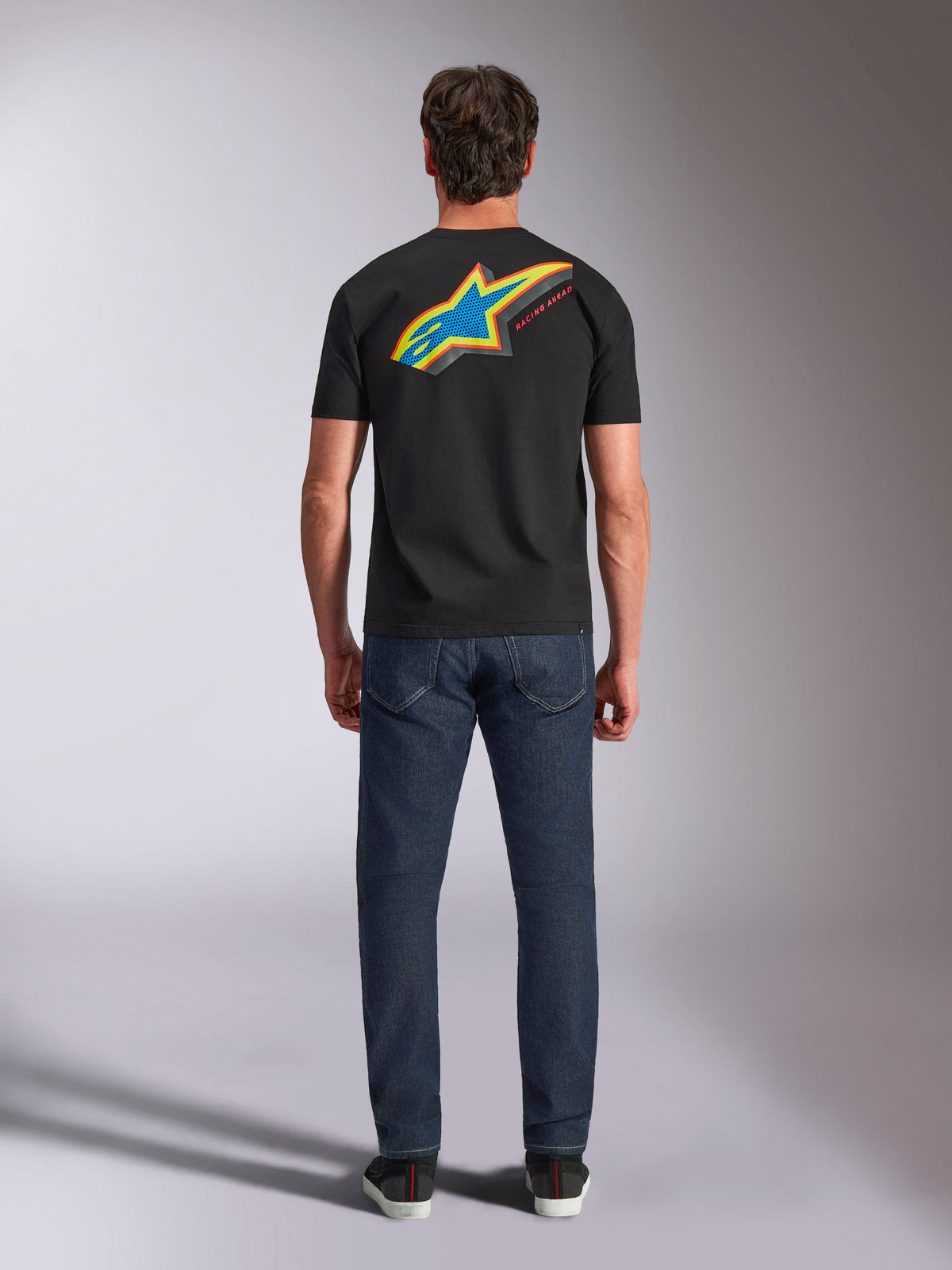 Alpinestars Angler CSF Tee, Short Sleeve T-Shirt, Black, Back view of model wearing shirt with large vibrant multi-color Astars star logo and 'RACING AHEAD' text, paired with dark blue jeans, casual motorsport streetwear
