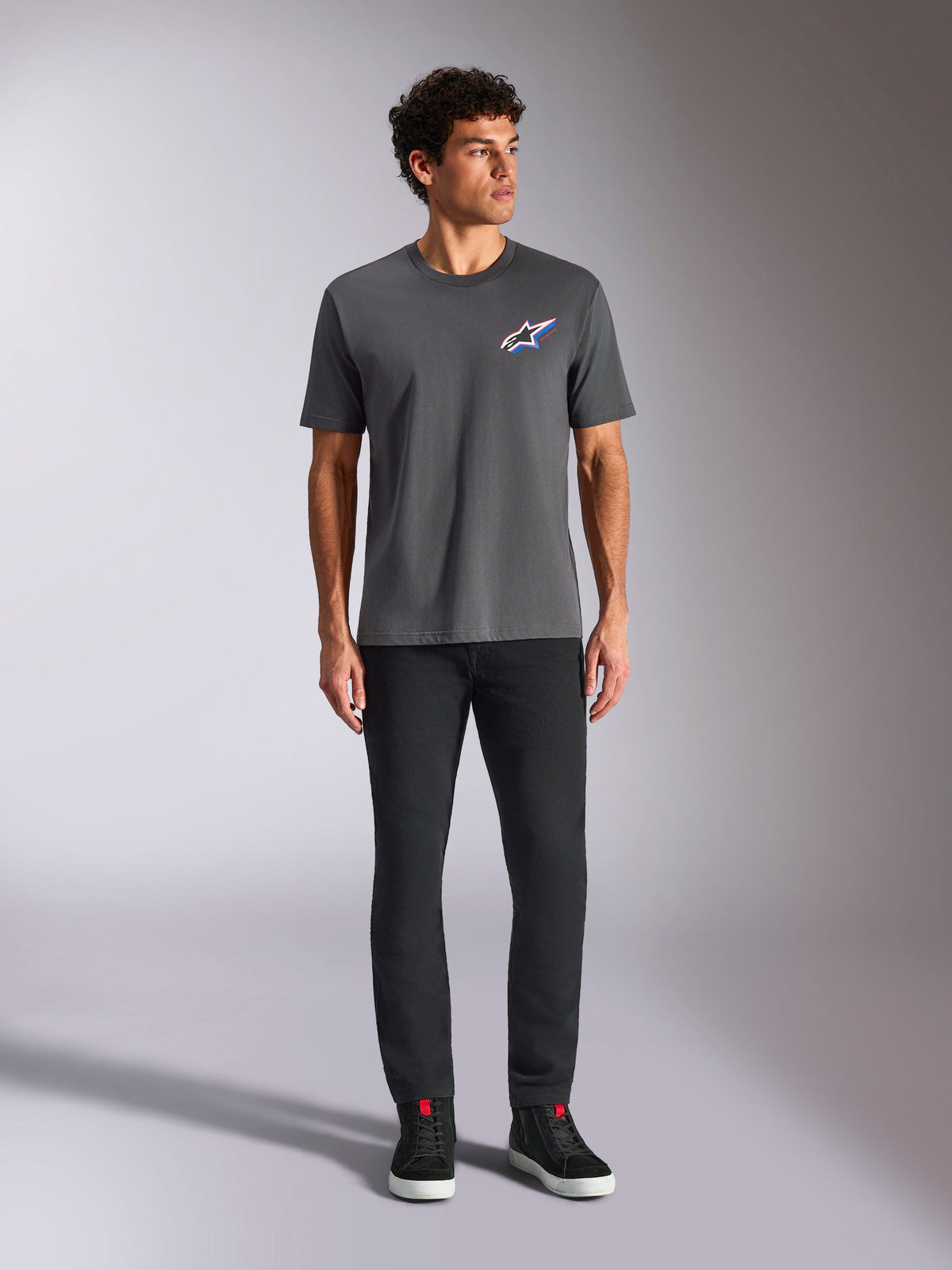 Alpinestars Angler CSF Tee, Kurzarm-T-Shirt, Anthrazit, mit einem lebendigen mehrfarbigen Astars-Logo auf der Brust, getragen von einem Model mit schwarzer Hose und Sneakern, Freizeit-Motorsport-Streetwear