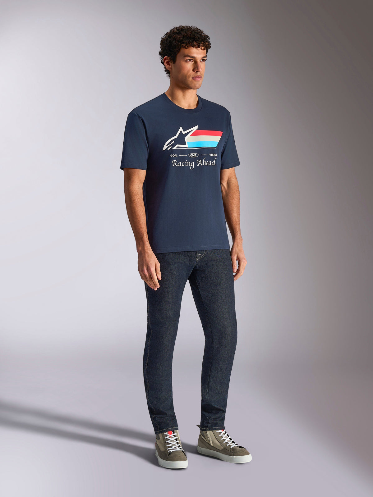 Alpinestars Trifecta CSF Tee, kurzärmeliges T-Shirt, Marineblau, mit Astars-Logo, dreifarbigen Streifen und „Racing Ahead“-Schriftzug, getragen von einem Model mit dunkler Jeans und Sneakern, lässige Motorsport-inspirierte Streetwear
