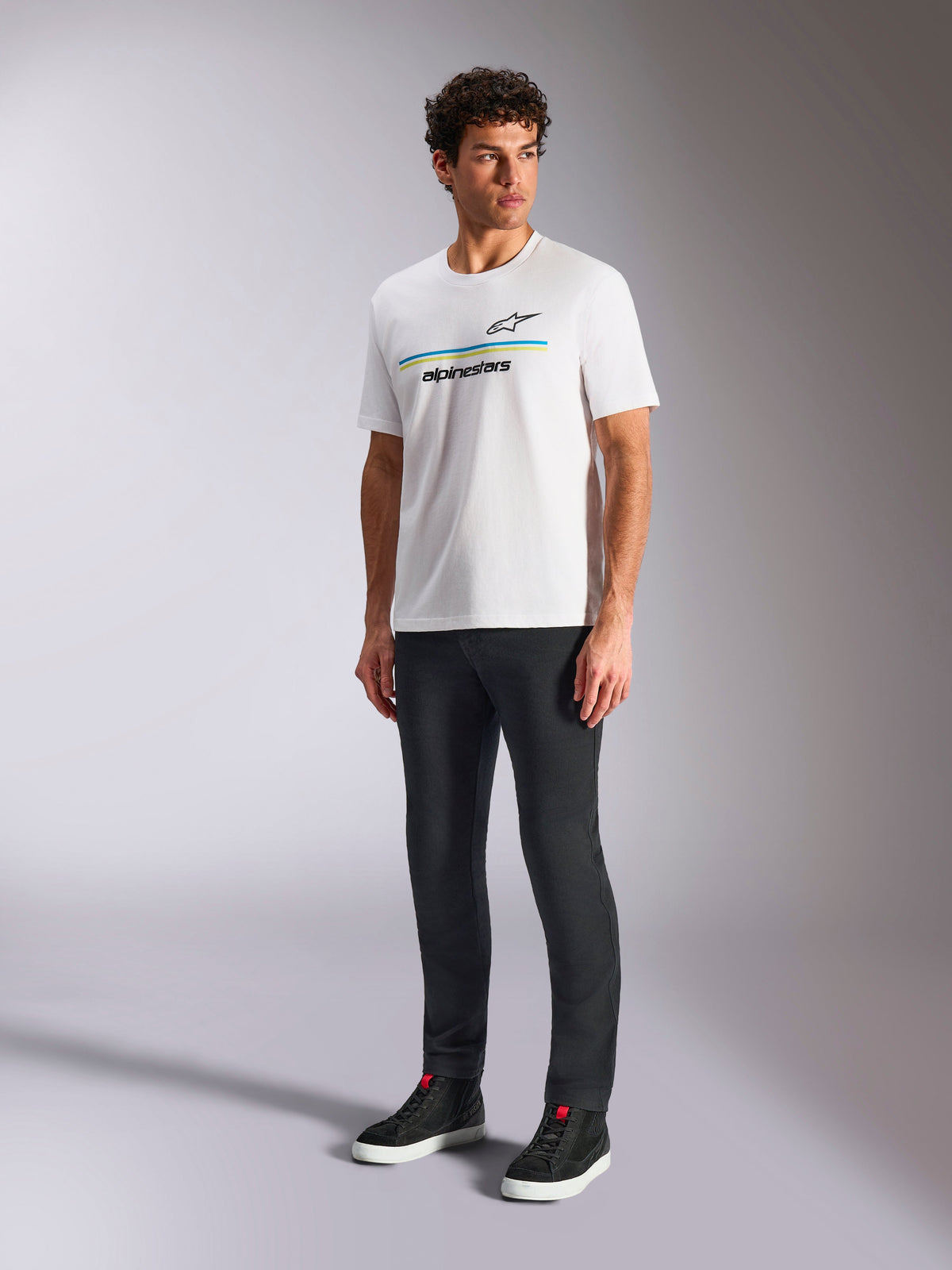 Alpinestars Lanes CSF Tee, t-shirt blanc à manches courtes, porté par un mannequin avec un pantalon slim noir et des baskets montantes noires, arborant des rayures horizontales bleues et jaunes avec le marquage Alpinestars sur la poitrine, streetwear décontracté inspiré du sport automobile