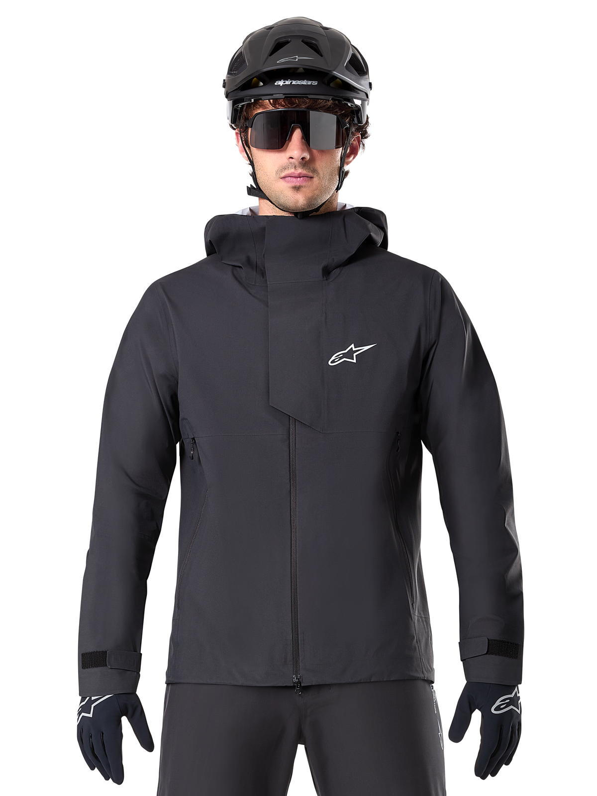 Alpinestars A-Dura Elite Waterproof Jacket, zwarte mountainbike jas, hoge kraag met capuchon, ritssluiting aan de voorzijde met beschermende borstflap en wit logo, gedragen met zwarte helm en fietshandschoenen, technische weersbescherming voor trailrijden