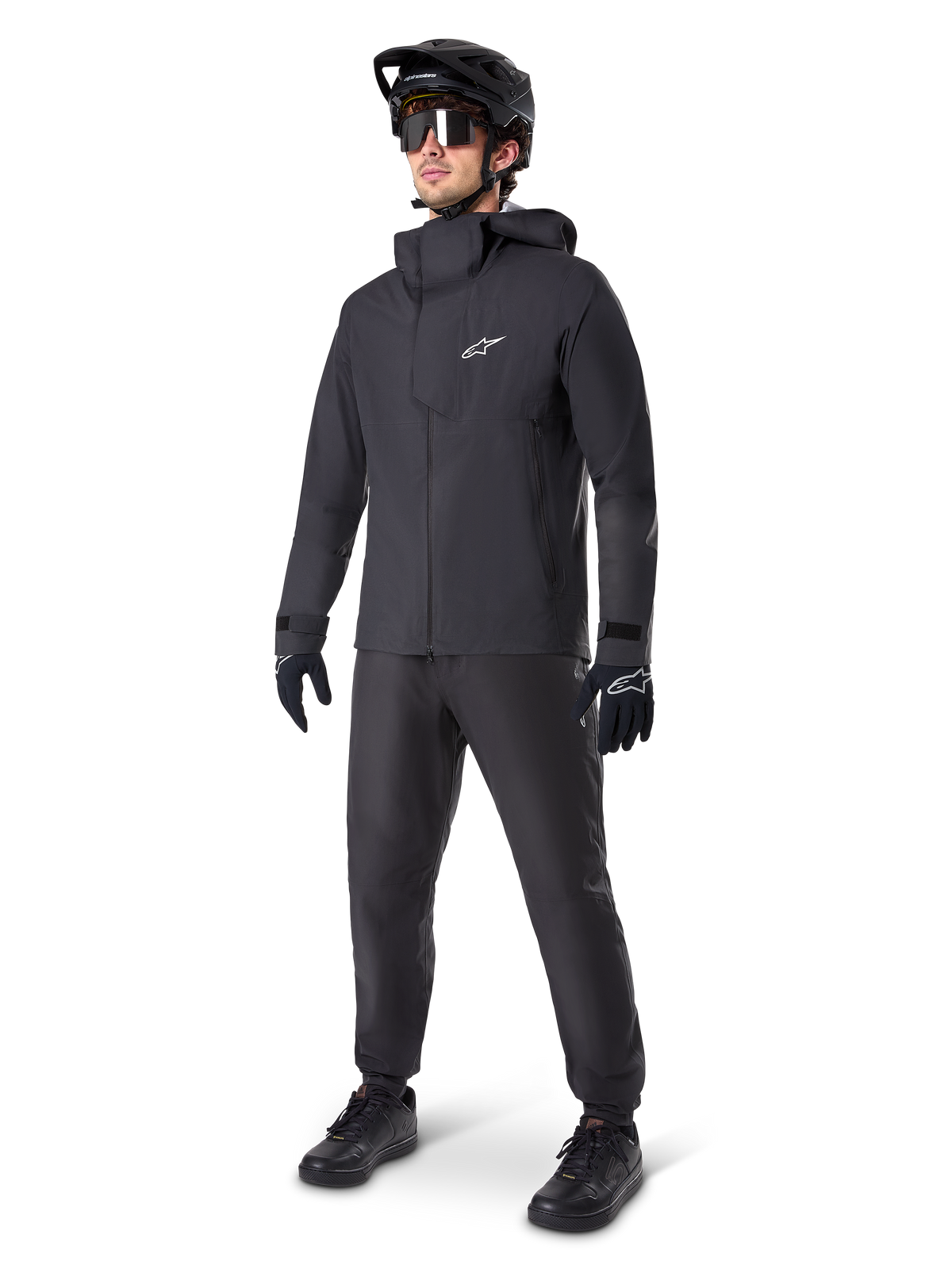 Alpinestars A-Dura Elite waterdichte jas in het zwart, gedragen door een mannelijke rider met bijpassende zwarte MTB-broek, handschoenen en helm. De jas heeft een technisch waterdicht ontwerp met een wit Astars-logo op de borst en een hoge beschermende kraag.
