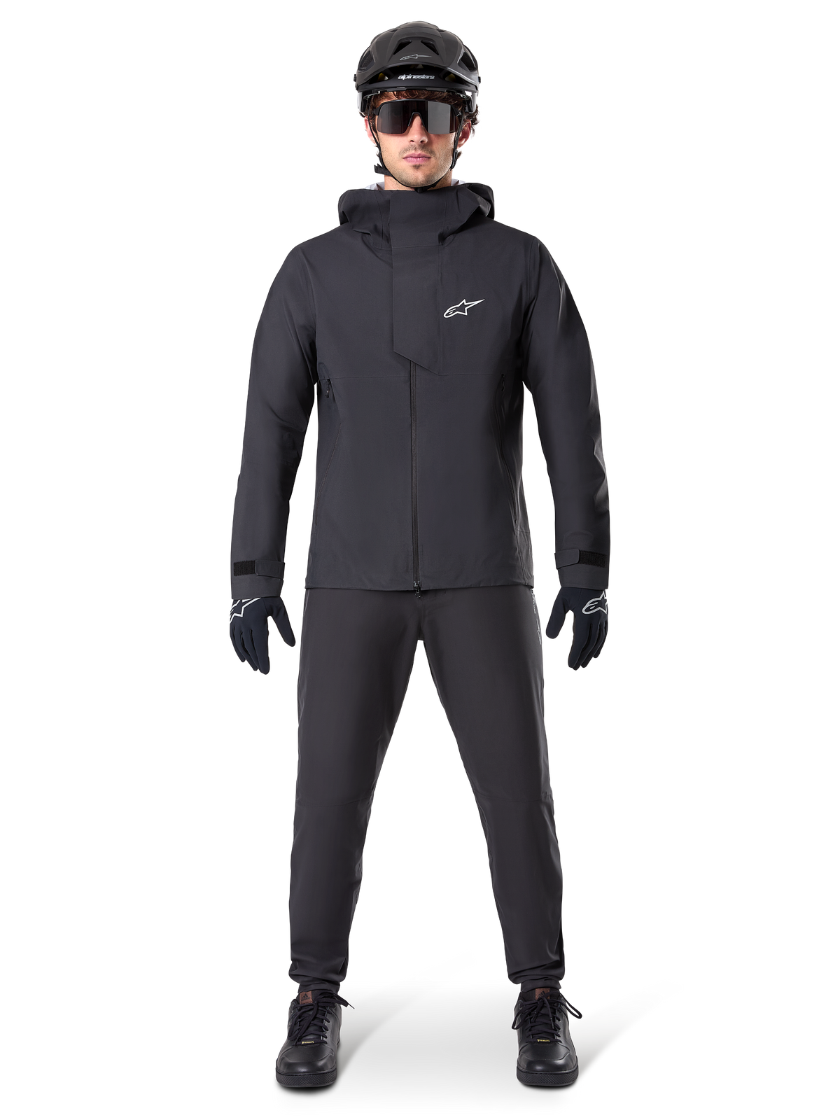 Alpinestars A-Dura Elite waterdichte jas, zwart, gedragen met bijpassende fietsbroek en handschoenen, zwarte helm en zonnebril, voorzien van een wit Astars-logo op de borst, hoogwaardig technisch ontwerp voor mountainbiken