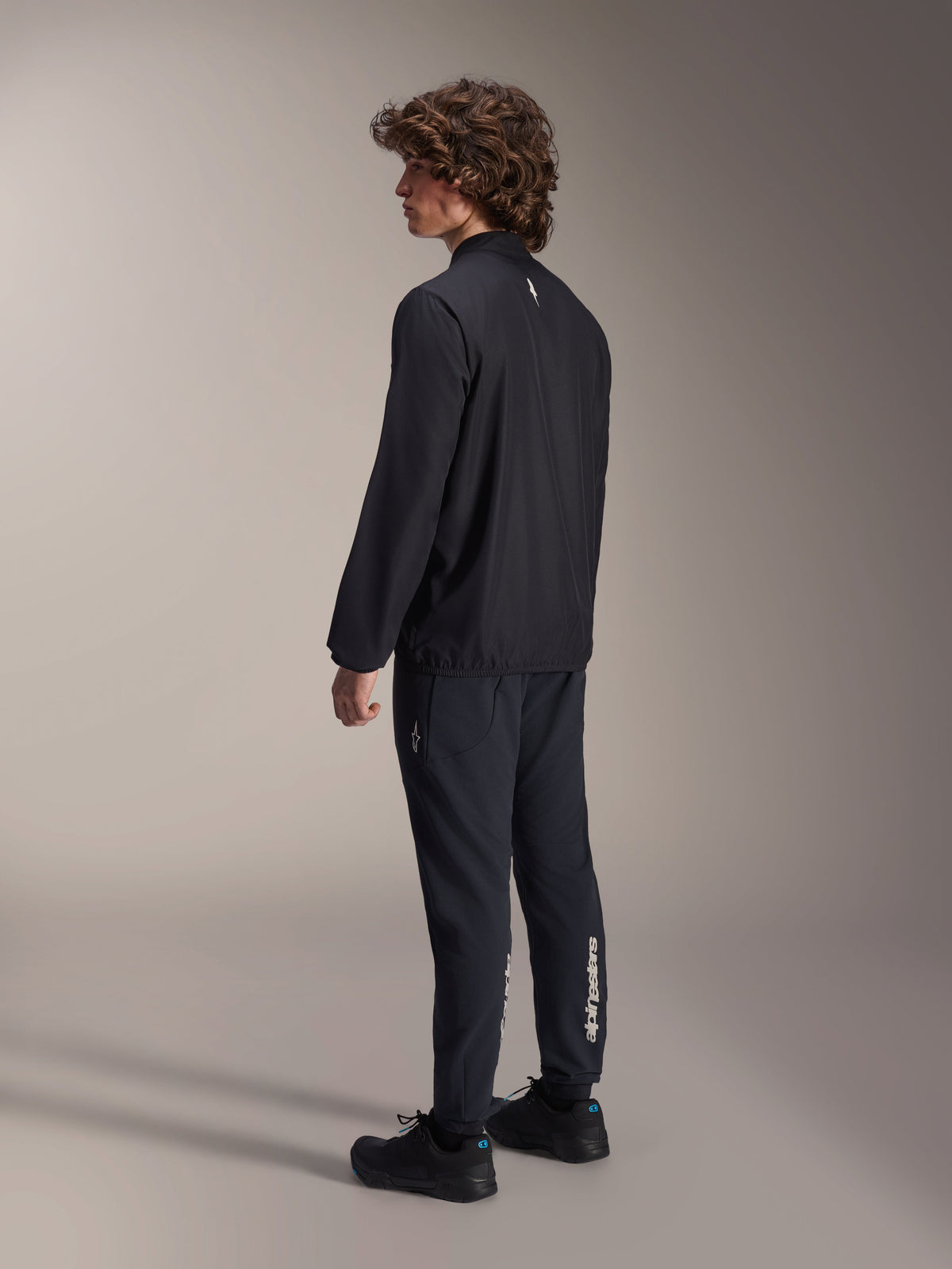 Alpinestars A-Aria Windbreaker-Jacke, Schwarz, leichte windabweisende Fahrradjacke, Dreiviertel-Rückansicht am Modell, mit kleinem weißem Logo im Nacken und elastischem Saum, kombiniert mit passender schwarzer Hose und Schuhen für einen performance-orientierten Look.