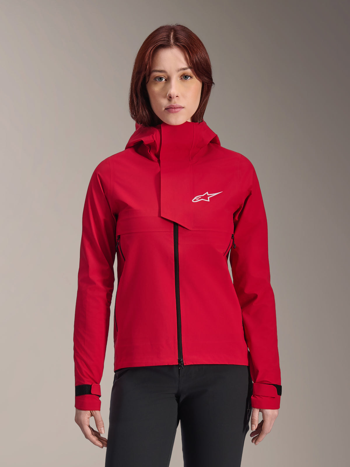 Veste imperméable Alpinestars Stella A-Dura Elite, veste de VTT pour femme, rouge fluo, design à capuche avec rabat frontal asymétrique, logo Alpinestars blanc sur la poitrine, poignets noirs ajustables, portée par un mannequin femme avec un pantalon noir