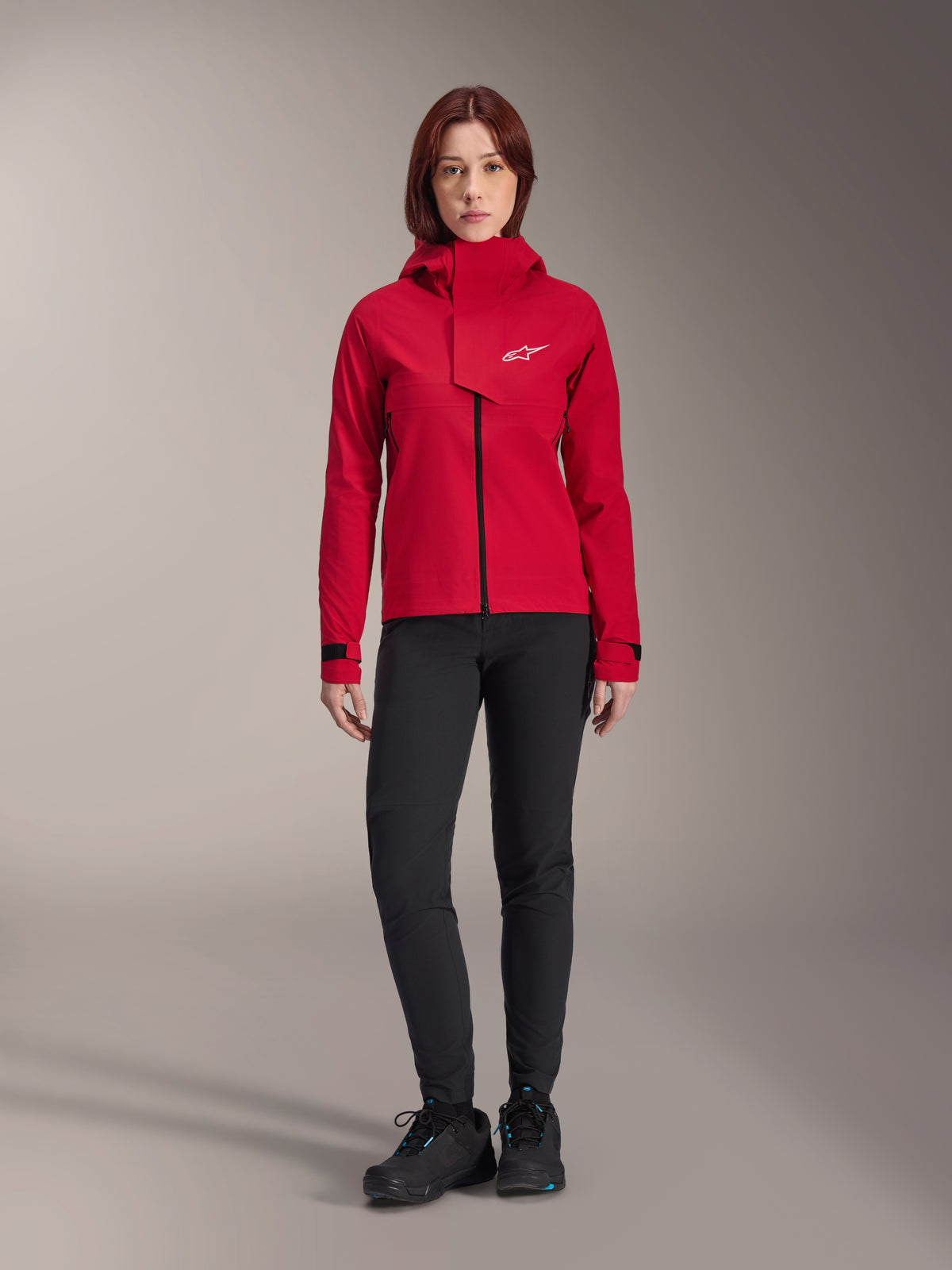Veste imperméable Stella A-Dura Elite Alpinestars, veste de VTT pour femme, Rouge Fluo, portée avec un pantalon et des chaussures noirs, dotée d'une capuche à col montant et d'un logo Alpinestars blanc, vêtement technique de VTT