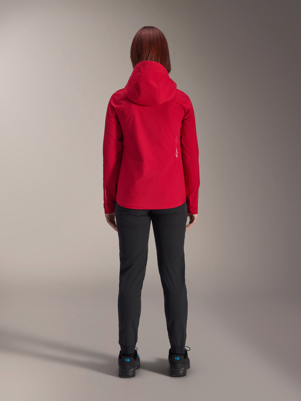 Vue arrière d'une femme portant la veste imperméable Alpinestars Stella A-Dura Elite en rouge fluo, associée à un pantalon et des chaussures noirs, dotée d'une capuche de protection et d'un ourlet arrière plus long pour le VTT.