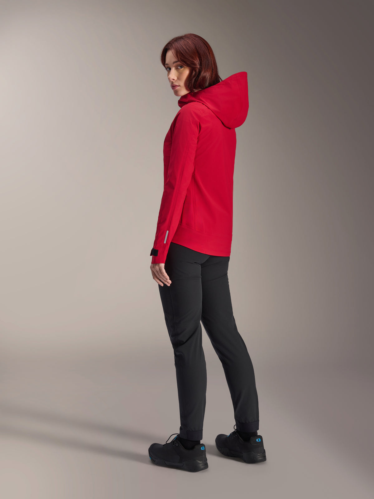 Femme portant la veste imperméable Alpinestars Stella A-Dura Elite en Rouge Fluo, associée à un pantalon de vélo technique noir et des chaussures noires, vue de trois-quarts arrière montrant la capuche réglable et l'ourlet arrière allongé, vêtement de performance pour le VTT