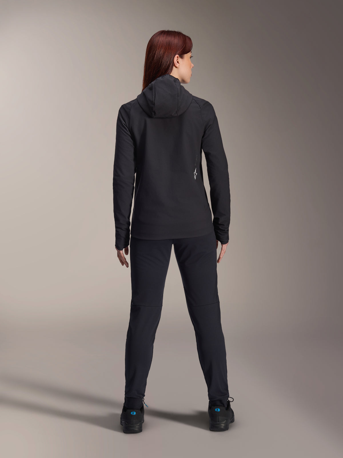 Femme portant la veste thermique Alpinestars Stella A-Dura, vue de dos, veste de cyclisme isolée noire avec capuche, pantalon et chaussures noirs assortis, avec un petit logo blanc dans le bas du dos