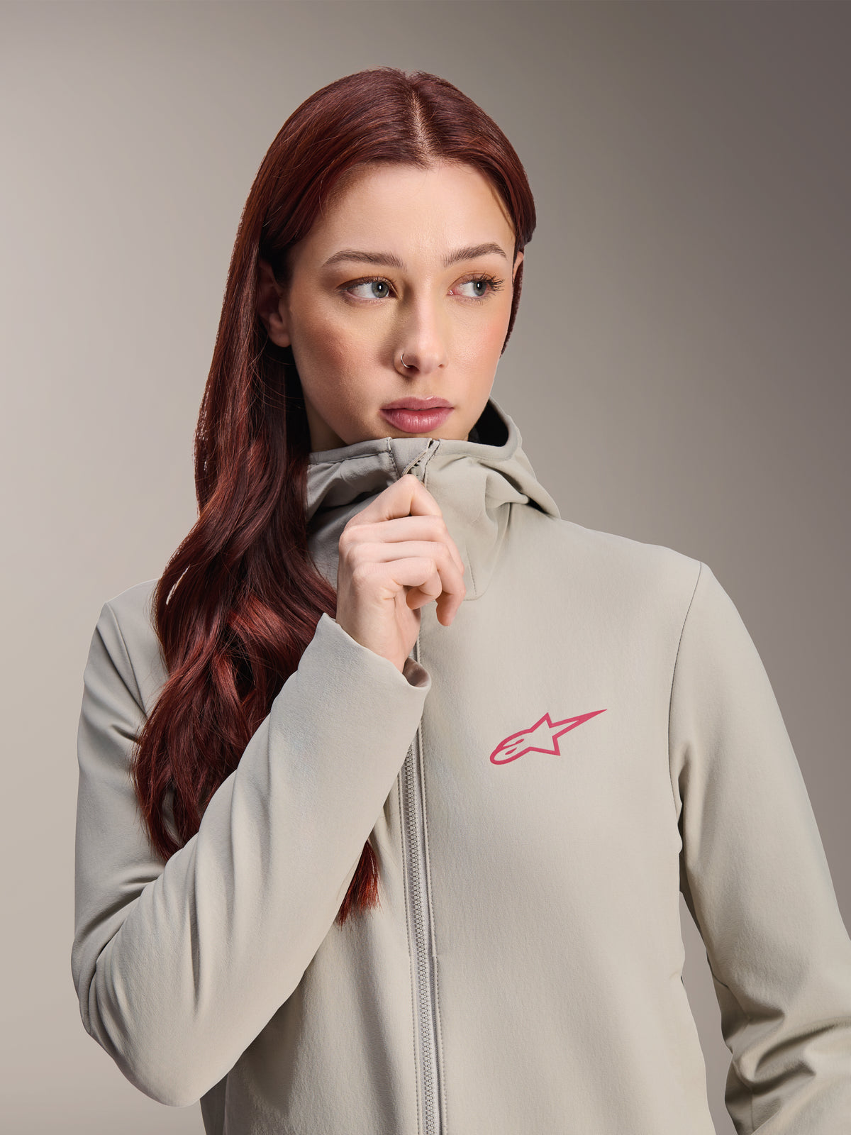Alpinestars Stella A-Dura Thermojas voor dames, titaniumgrijs met een roze Alpinestars-sterlogo, model met lang rood haar die de jas met capuchon draagt en dichtritst, lichtgewicht geïsoleerde fietskleding voor koud weer.