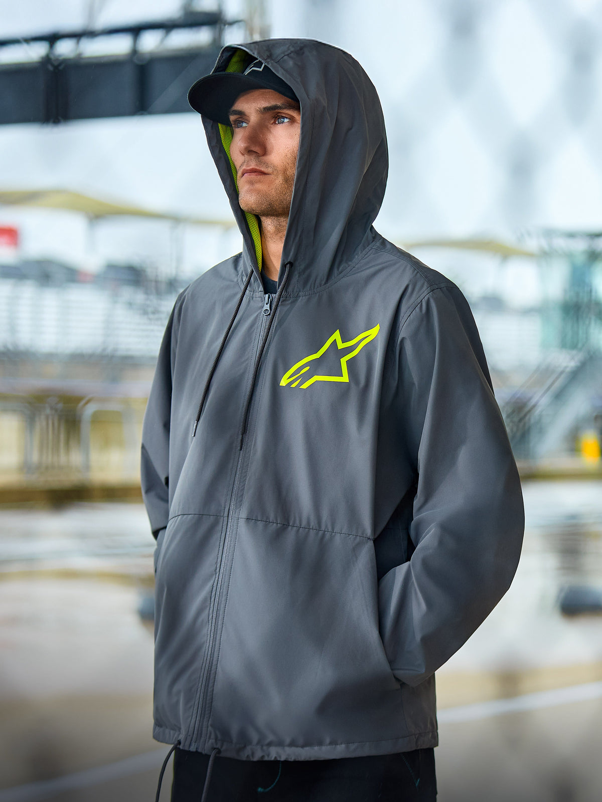 Alpinestars Speeder Chromium Windbreak Jacke, anthrazitgraue Lifestyle-Jacke mit neongelben Akzenten, Kapuzendesign mit gelbem Innenfutter und Brustlogo, lässige Motorsport-Streetwear, getragen von einem Model im Freien