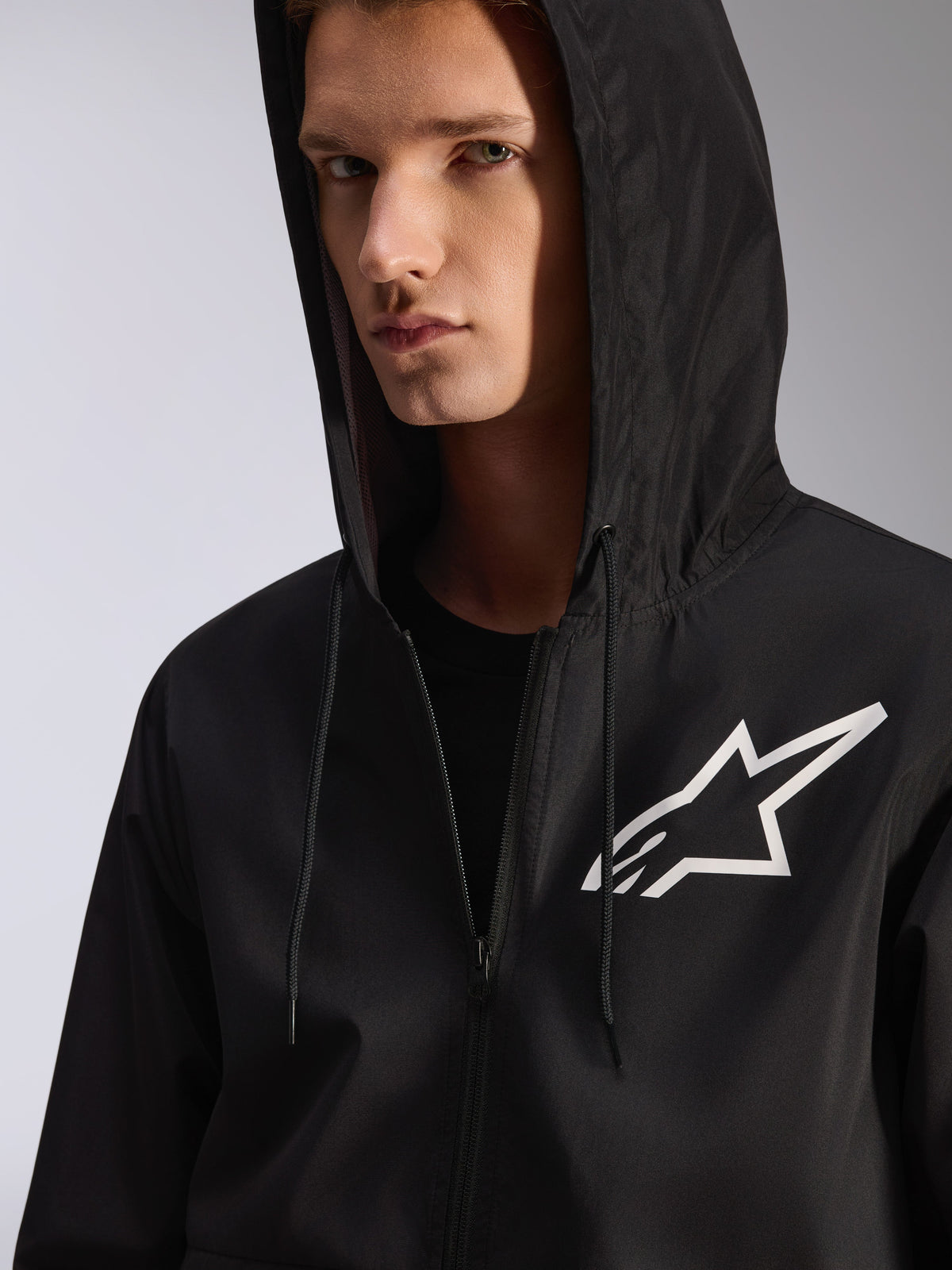 Alpinestars Speeder Chromium Windbreaker-Jacke, Schwarz, Nahaufnahme einer Windjacke mit Kapuze und weißem Stern-Logo auf der Brust, verfügt über eine Kapuze mit Kordelzug, leichtes Textilmaterial, lässige, vom Motorsport inspirierte Streetwear