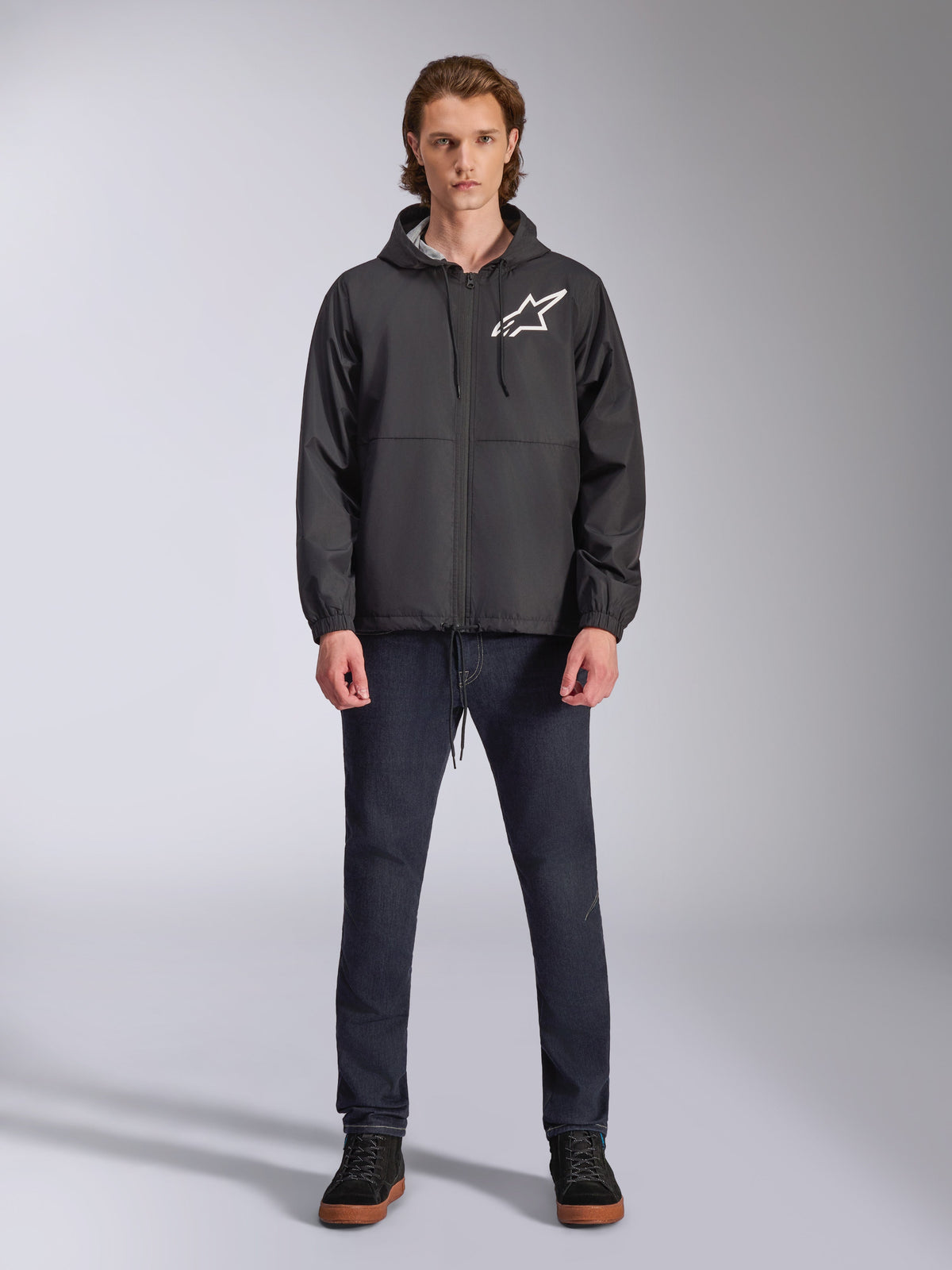 Alpinestars Speeder Chromium Windbreaker-Jacke, Schwarz mit weißem Astars-Logo, getragen von einem Model mit dunkelblauen Jeans und schwarzen Sneakern, mit Kapuzendesign und Frontreißverschluss, lässige vom Motorsport inspirierte Streetwear