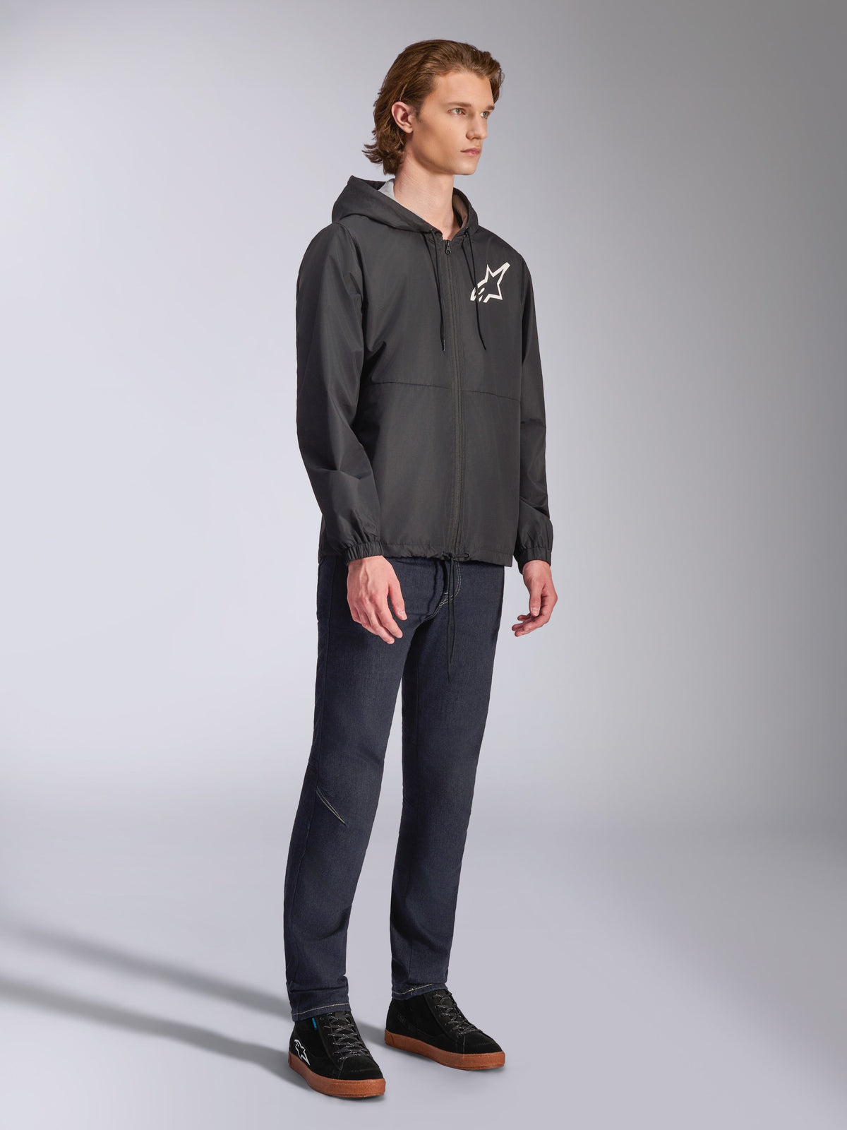 Alpinestars Speeder Chromium Windbreak Jacket, Schwarz, Kapuzen-Windbreaker mit Frontreißverschluss, weißes Alpinestars Stern-Logo auf der linken Brust, lässige Motorsport-Streetwear, kombiniert mit dunkler Hose und schwarzen Sneakern in einem Studio-Setting.