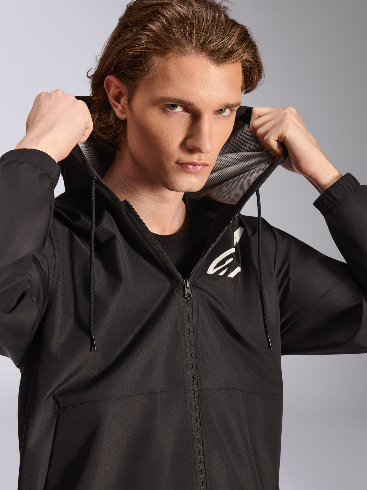 Alpinestars Speeder Chromium Windbreaker-Jacke, Schwarz, getragen von einem männlichen Modell, das die Kapuze hält, weißes Logo auf der Brust, Mesh-Kapuzenfutter, durchgehender Frontreißverschluss, lässige Streetwear im Motorsport-Stil