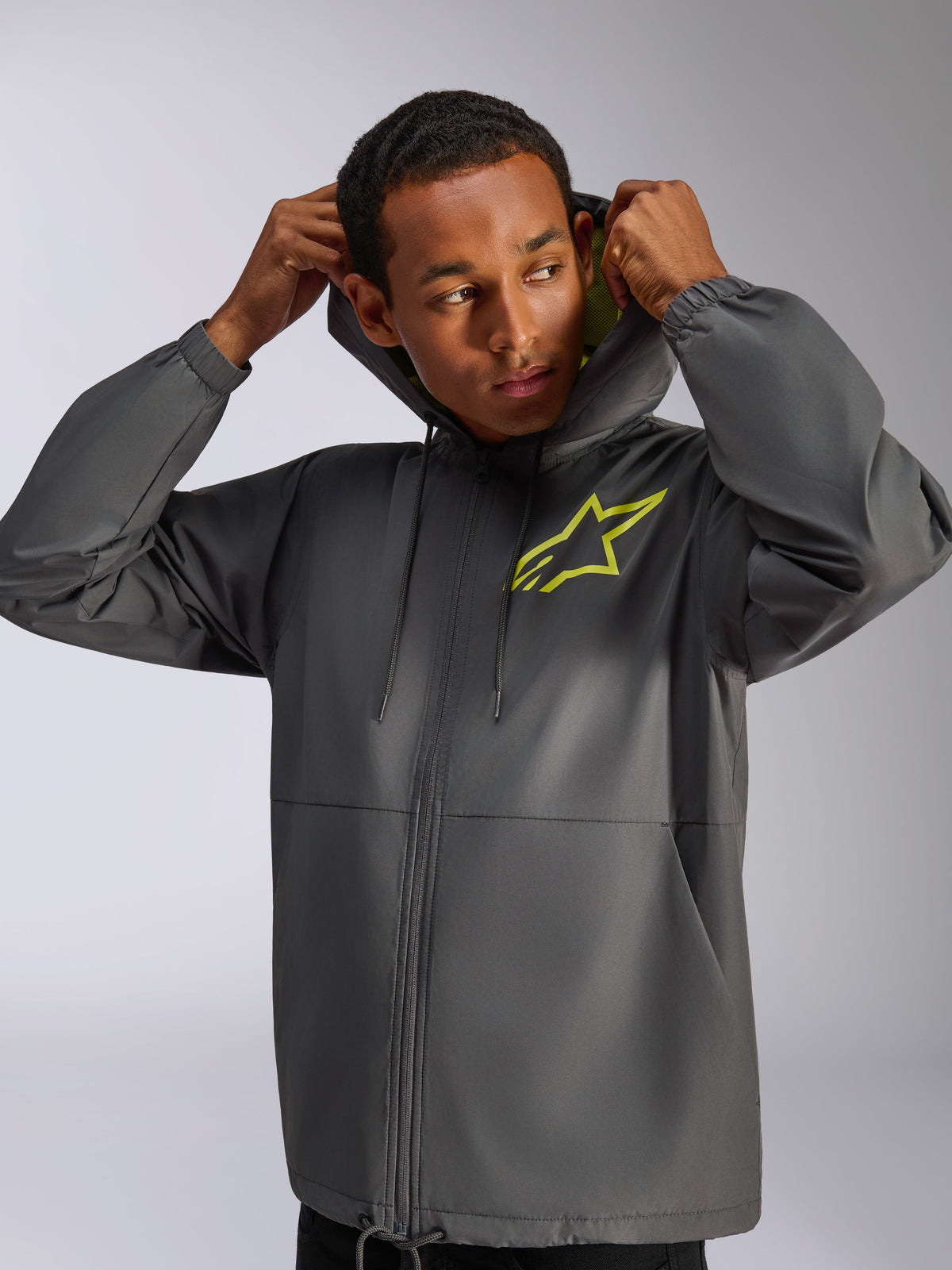 Alpinestars Speeder Chromium Windbreaker-Jacke, Anthrazitgrau und Neongelb, männliches Model trägt Kapuzen-Windbreaker mit Kordelzug, neongelbes Alpinestars-Logo auf der Brust und gelbes Kapuzenfutter, lässige vom Motorsport inspirierte Streetwear