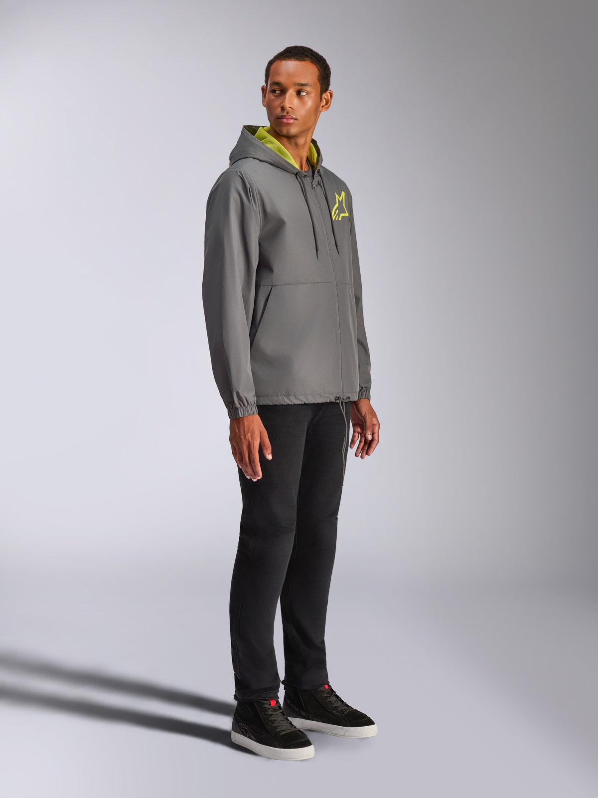 Alpinestars Speeder Chromium Windbreaker-Jacke, Anthrazit mit neongelben Akzenten, männliches Model trägt Windbreaker mit Kapuze, gelbes Stern-Logo auf der Brust, kombiniert mit schwarzer Hose und schwarzen Sneakern, lässige vom Motorsport inspirierte Streetwear