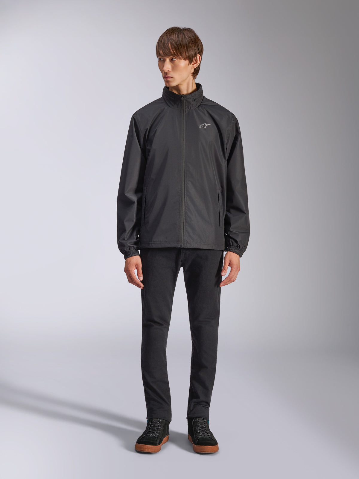 Alpinestars Overhaul Racer Jacket, Black Men's Rain Shell, full-length view on a model paired with black pants and casual sneakers, featuring a small white logo on the chest, water-resistant fabric, and casual motorsport-inspired design