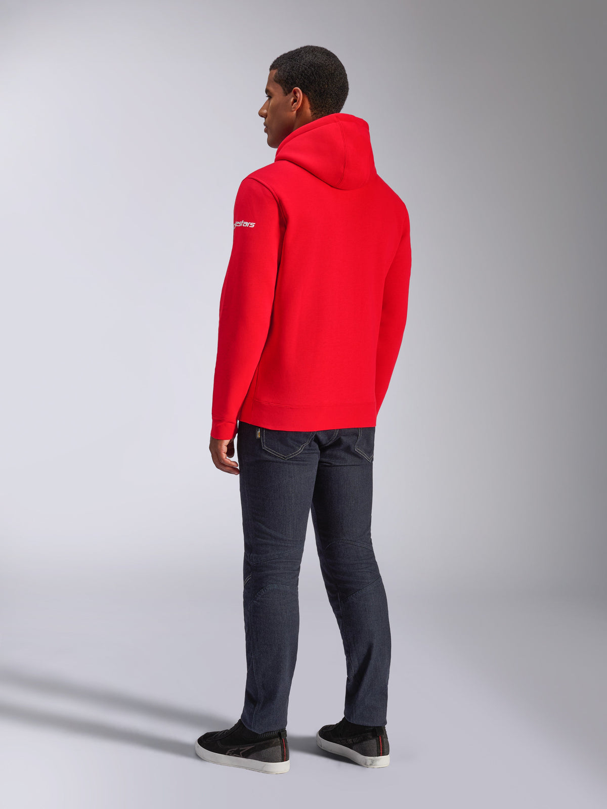 Alpinestars Frontal Hoodie, rode pullover fleece hoodie, achteraanzicht met wit woordmerklogo op de mouw, gedragen met donkere denim broek en zwarte schoenen, casual motorsport streetwear