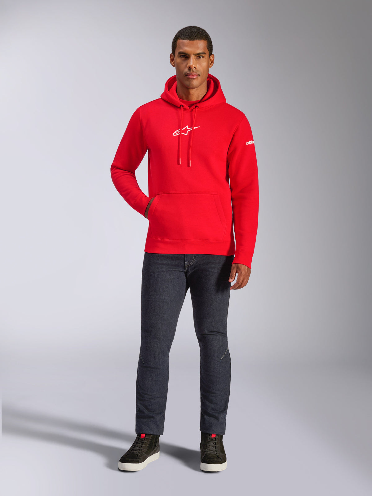 Mannelijk model draagt de rode Alpinestars Frontal Hoodie, een pullover fleece hoodie met wit Astars-logo op de borst, kangoeroezak en capuchon met trekkoord, gecombineerd met een donkergrijze jeans en zwarte hoge sneakers, casual motorsport streetwear