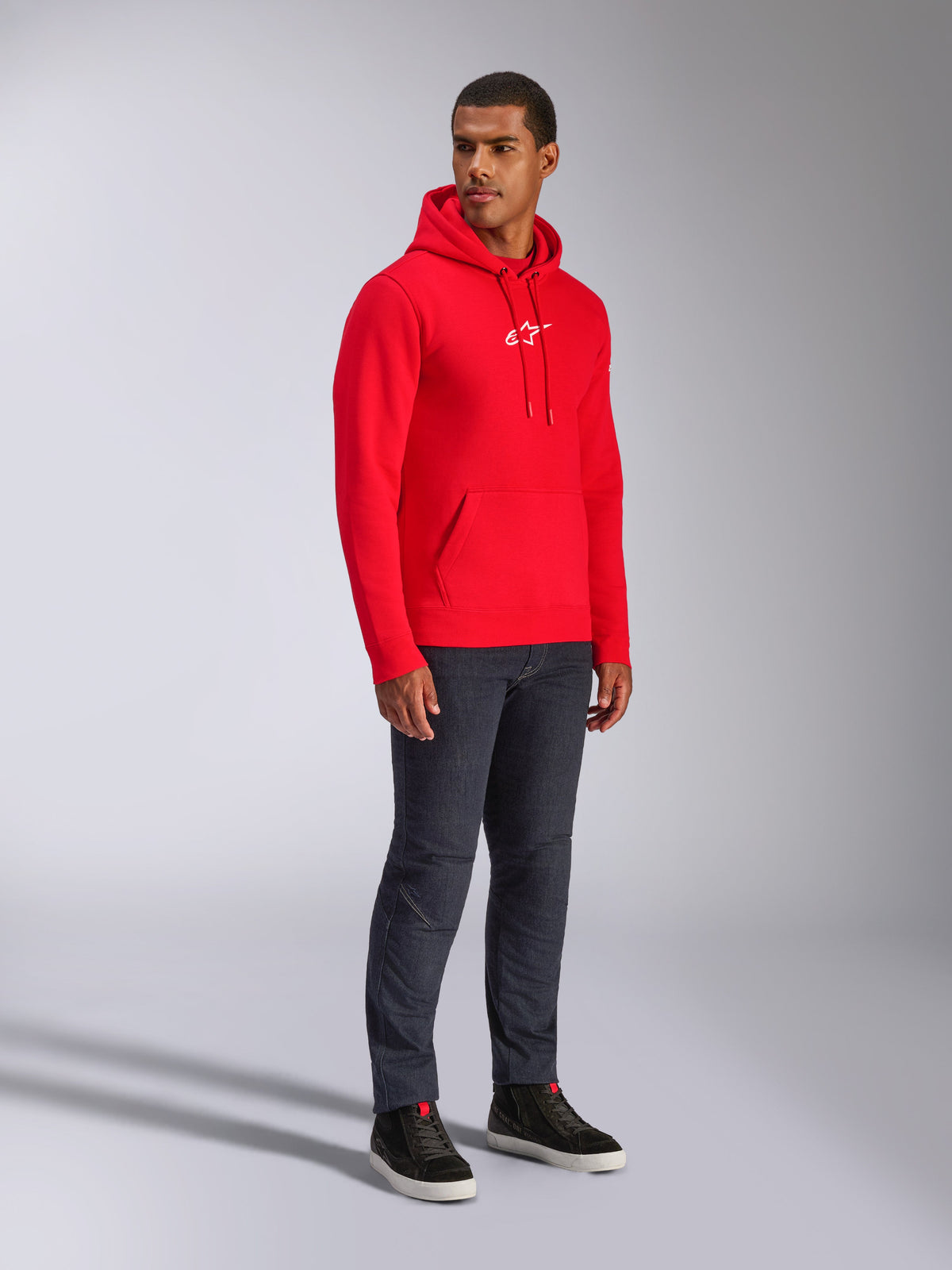 Alpinestars Frontal Hoodie, rood, mannelijk model in pullover fleece hoodie met wit A-star logo op de borst, kangoeroezak en capuchon met trekkoord, gecombineerd met donkere denim jeans, casual op motorsport geïnspireerde streetwear