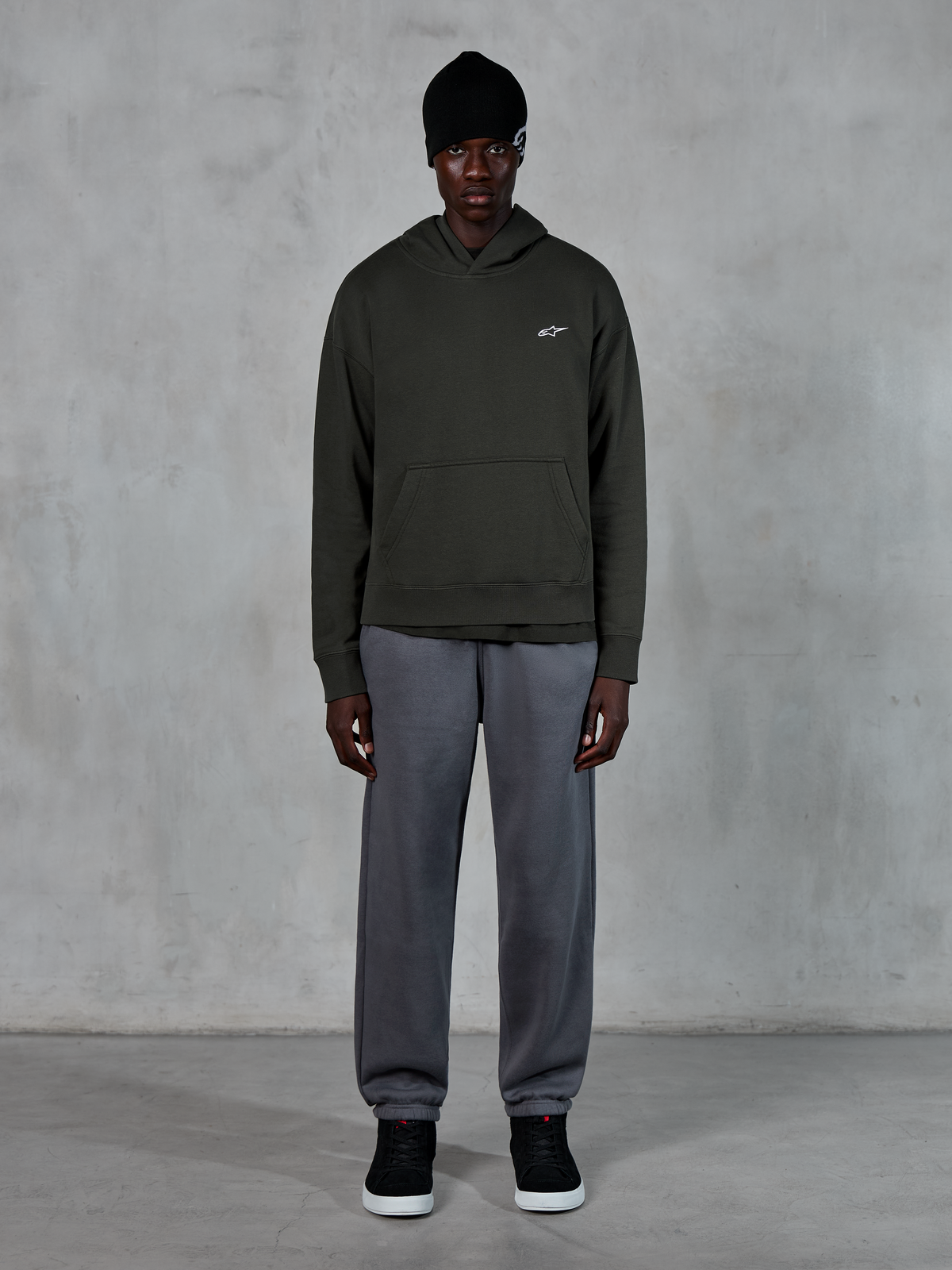 Model wearing Alpinestars Essentials Ageless Hoodie, casual streetwear, Off Black with small white logo on chest, paired with gray joggers, black beanie, and black sneakers, minimalist motorsport-inspired design