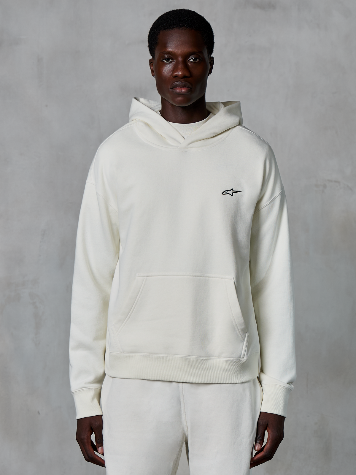 Alpinestars Essentials Ageless Hoodie, off-white, gedragen door mannelijk model, klein zwart logo op de borst, kangoeroezak aan de voorzijde, lange mouwen, casual streetwear geïnspireerd op de motorsport