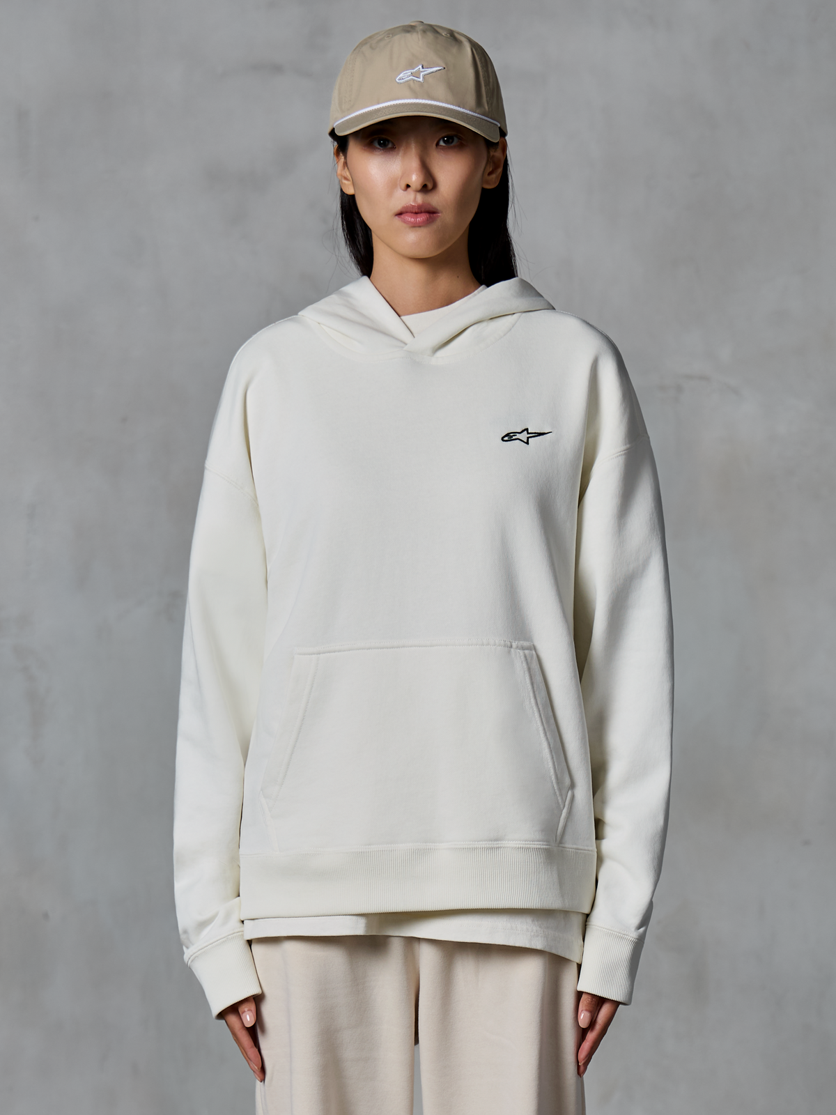 Alpinestars Essentials Ageless Hoodie, off-white pullover fleece hoodie, vrouwelijk model met beige pet en bijpassende crème joggingbroek, klein zwart Alpinestars-logo op de borst, kangoeroezak aan de voorkant, ontspannen pasvorm, casual motorsport-geïnspireerde streetwear