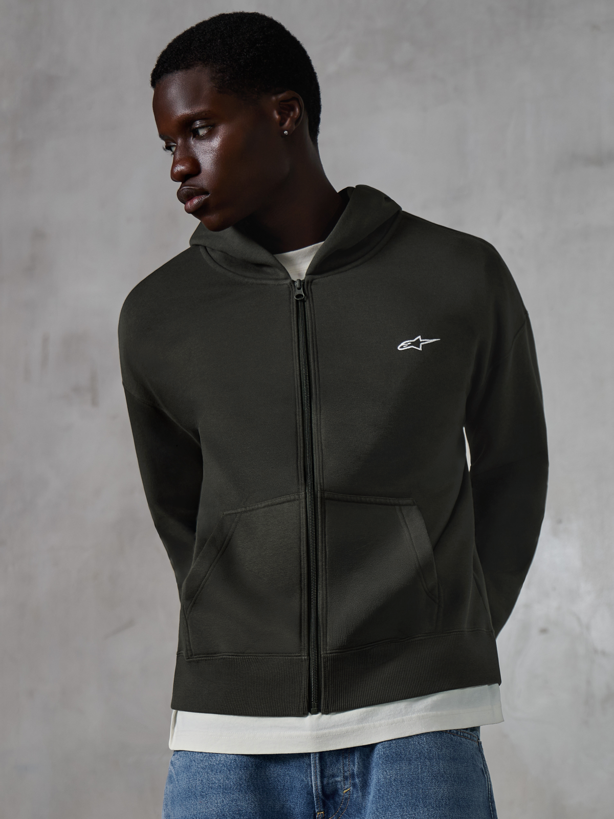 Hoodie zippé Essentials Ageless Alpinestars, streetwear décontracté noir délavé (Off Black), polaire entièrement zippée avec un petit logo Alpinestars blanc sur la poitrine, porté par un mannequin avec un jean bleu, coupe décontractée avec poches avant et finition délavée.