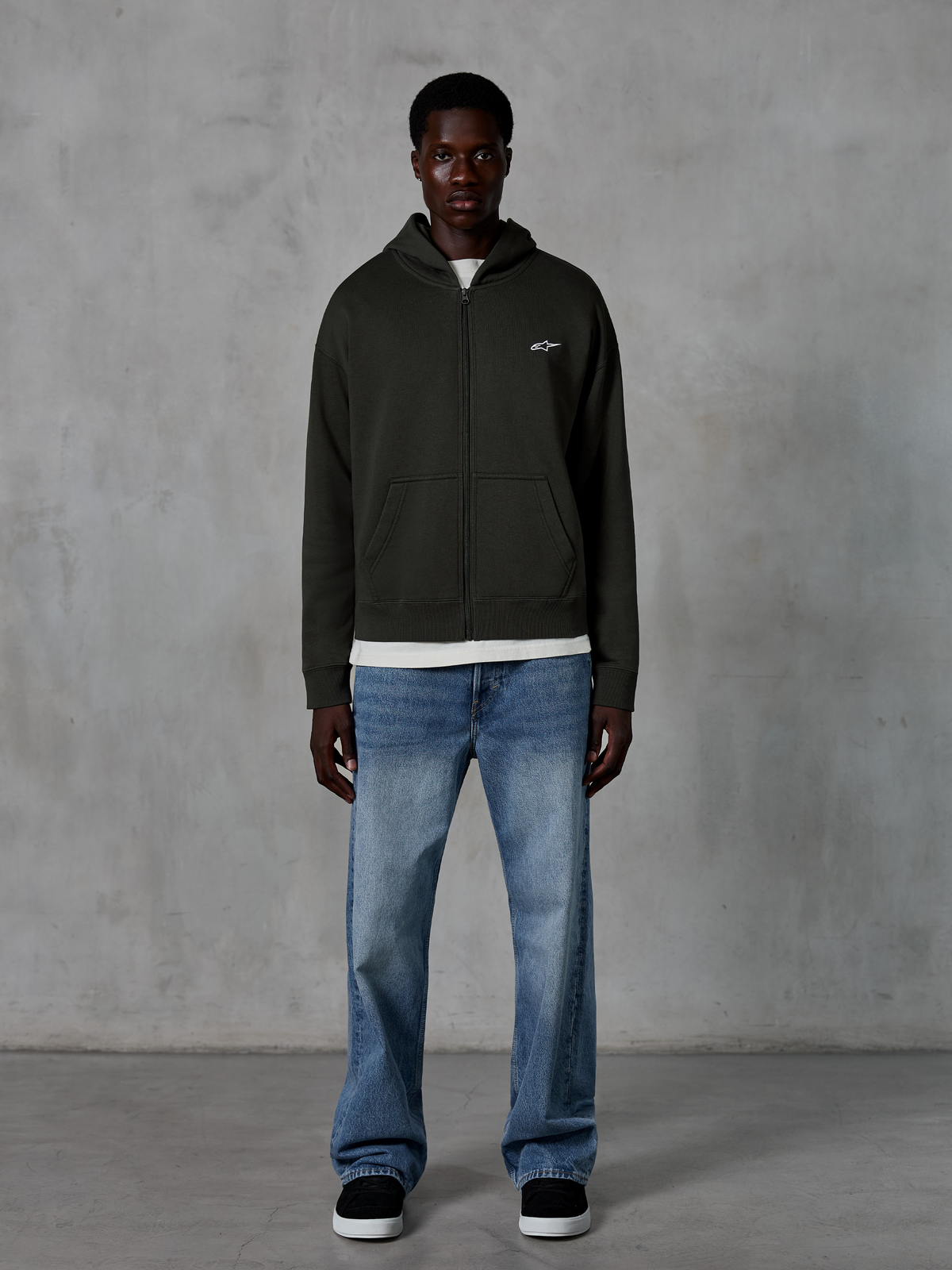 Sweat à capuche zippé Alpinestars Essentials Ageless, hoodie entièrement zippé, noir, avec un petit logo Alpinestars blanc sur la poitrine, porté par un mannequin avec un t-shirt blanc et un jean bleu, coupe décontractée, style streetwear décontracté motorsport