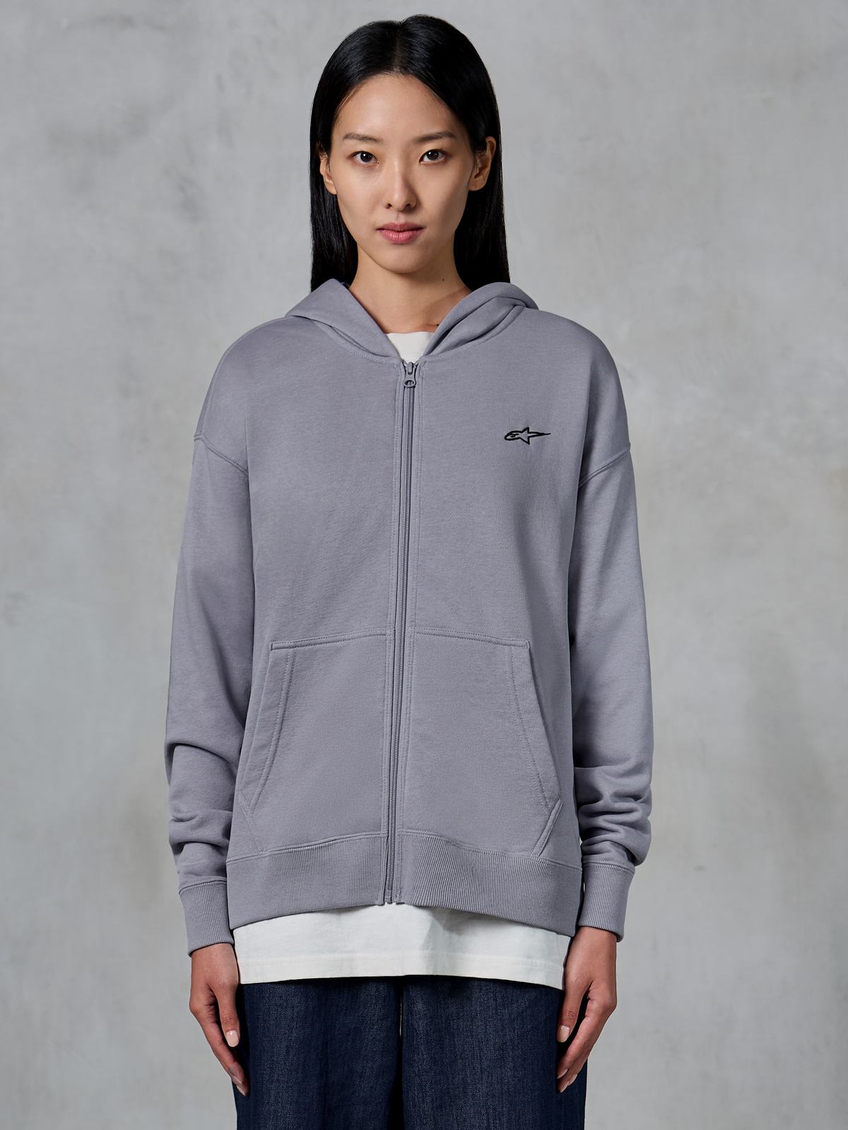 Sweat à capuche zippé Alpinestars Essentials Ageless, Silver, streetwear décontracté, sweat en polaire à fermeture éclair intégrale avec petit logo Alpinestars noir sur la poitrine, poche kangourou divisée, porté par un mannequin sur un t-shirt blanc et un jean foncé