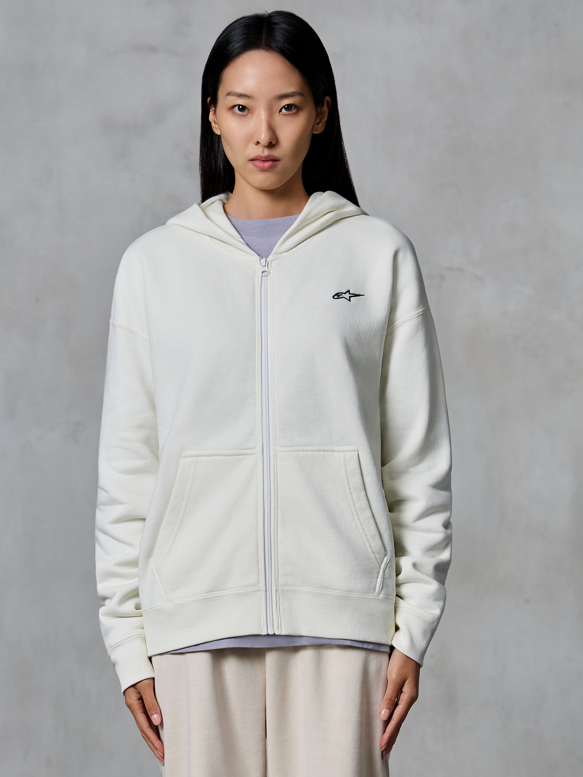 Sweat à capuche zippé Alpinestars Essentials Ageless, blanc cassé, sweat en polaire zippé porté par un mannequin, avec un petit logo Astars noir sur la poitrine, coupe décontractée, vêtement lifestyle décontracté inspiré du sport automobile