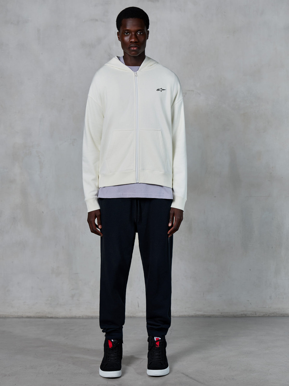 Alpinestars Essentials Ageless Zip Hoodie, sweat à capuche zippé blanc cassé, coupe décontractée avec petit logo noir sur la poitrine, porté avec un pantalon de jogging noir et des baskets, style streetwear décontracté inspiré du sport automobile