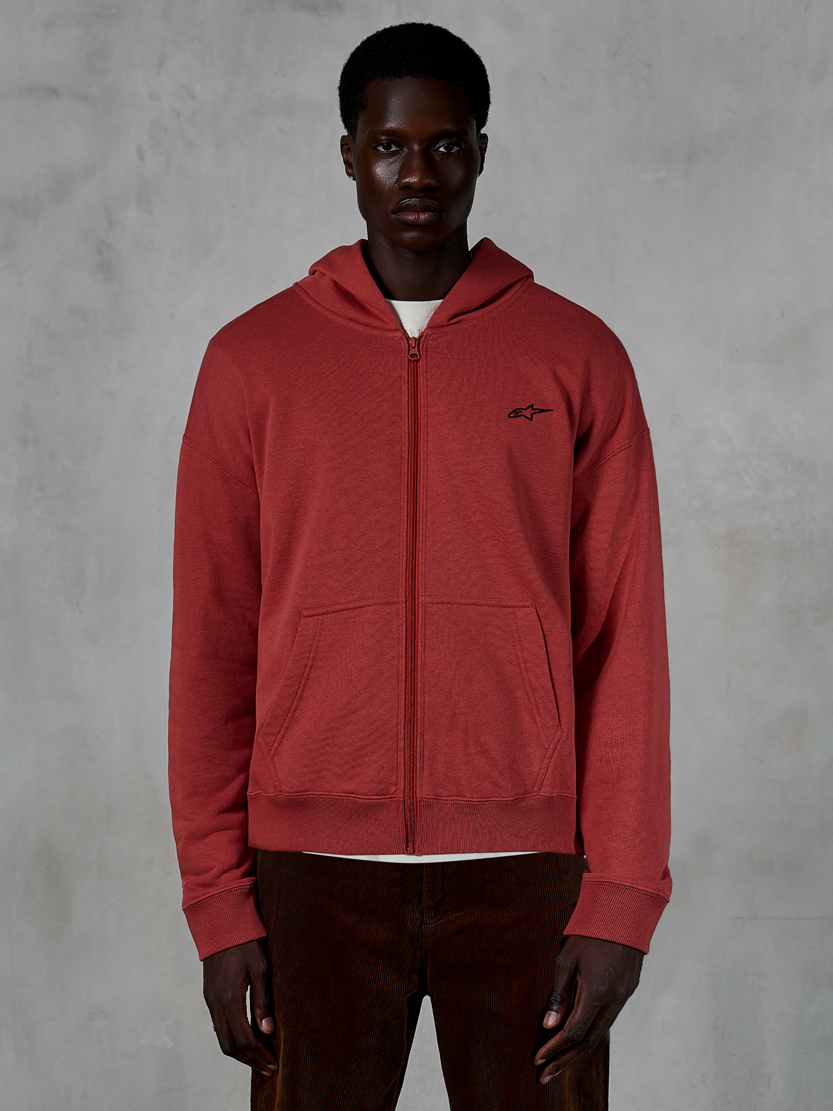 Alpinestars Essentials Ageless Zip Hoodie, ziegelrotes Kapuzensweatshirt mit durchgehendem Reißverschluss, getragen von einem Model mit brauner Cordhose, mit einem kleinen schwarzen Logo auf der Brust, lässige Streetwear in entspannter Passform