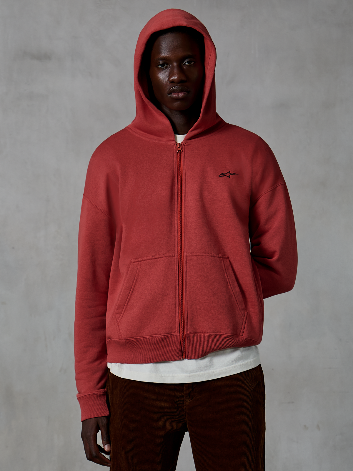 Alpinestars Essentials Ageless Zip Hoodie, lässige Sportbekleidung in Ziegelrot, Design mit durchgehendem Reißverschluss und aufgesetzter Kapuze, kleines schwarzes Logo auf der Brust, getragen von einem Model mit weißem T-Shirt und brauner Cordhose vor grauem Hintergrund.