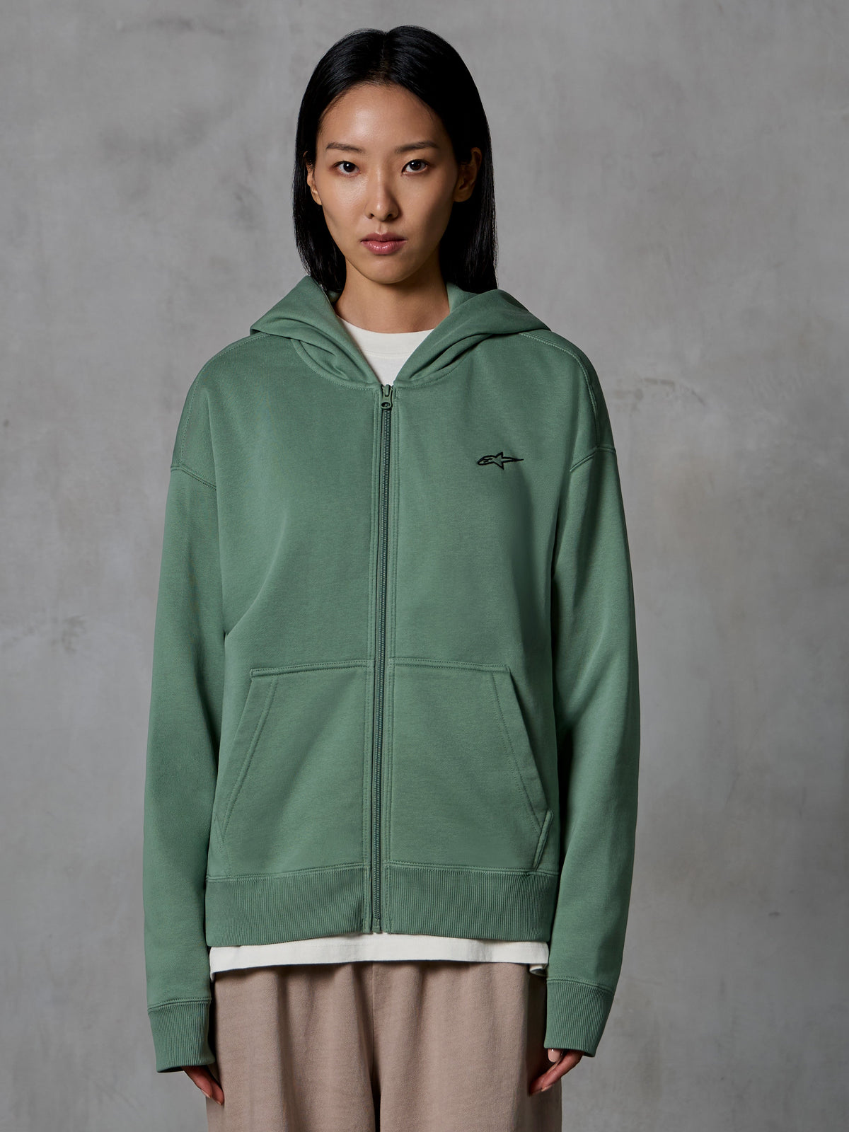 Alpinestars Essentials Ageless Zip Hoodie, Kaktusgrün, legere Streetwear getragen von einem weiblichen Model, Fleece-Sweatshirt mit Reißverschluss und kleinem schwarzen Logo auf der Brust, Fronttaschen, entspannte Passform für den Alltag