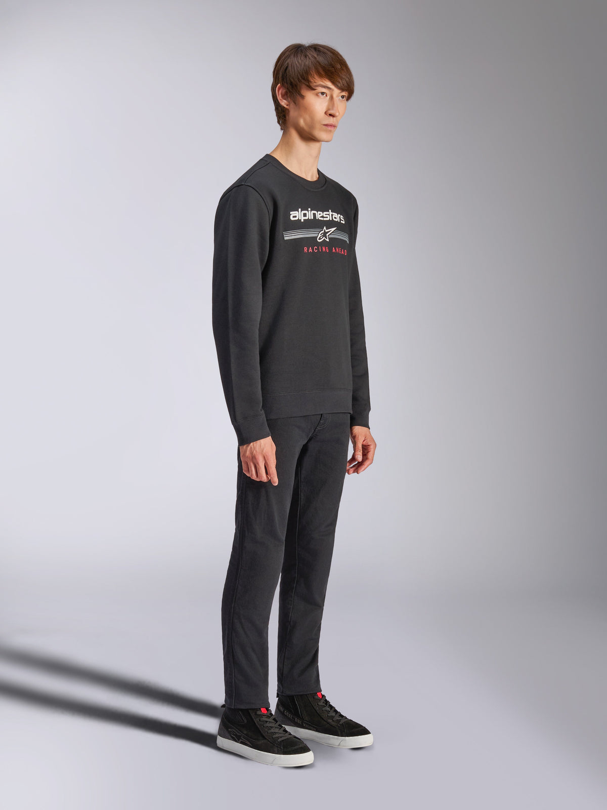 Alpinestars Bettern Crew Sweatshirt, schwarzer Rundhals-Pullover, weißes Alpinestars-Logo mit „RACING AHEAD“ in Rot auf der Brust, kombiniert mit schwarzer Hose und Sneakern, lässige, vom Motorsport inspirierte Streetwear