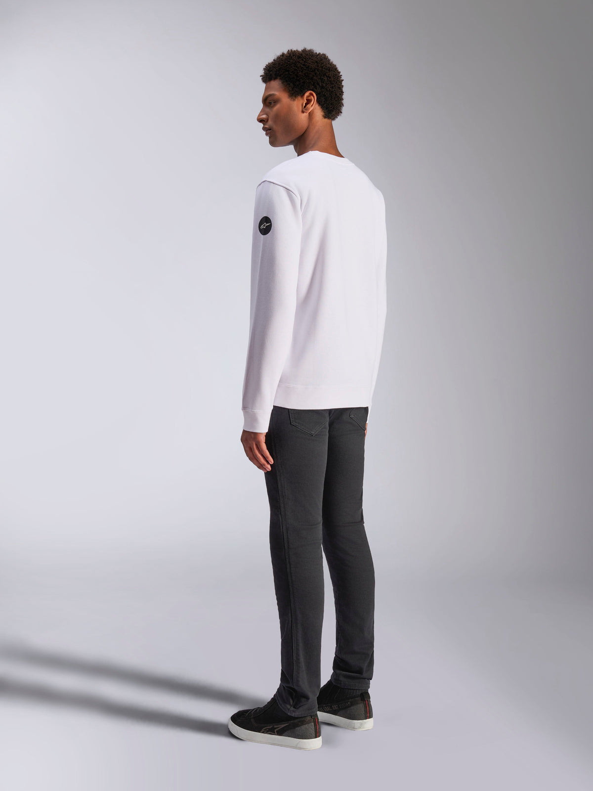 Alpinestars Bettern Crew, sweat-shirt à col rond blanc, vue de dos et de côté, logo circulaire noir sur la manche, porté avec un pantalon gris foncé et des baskets noires, style streetwear motorsport décontracté