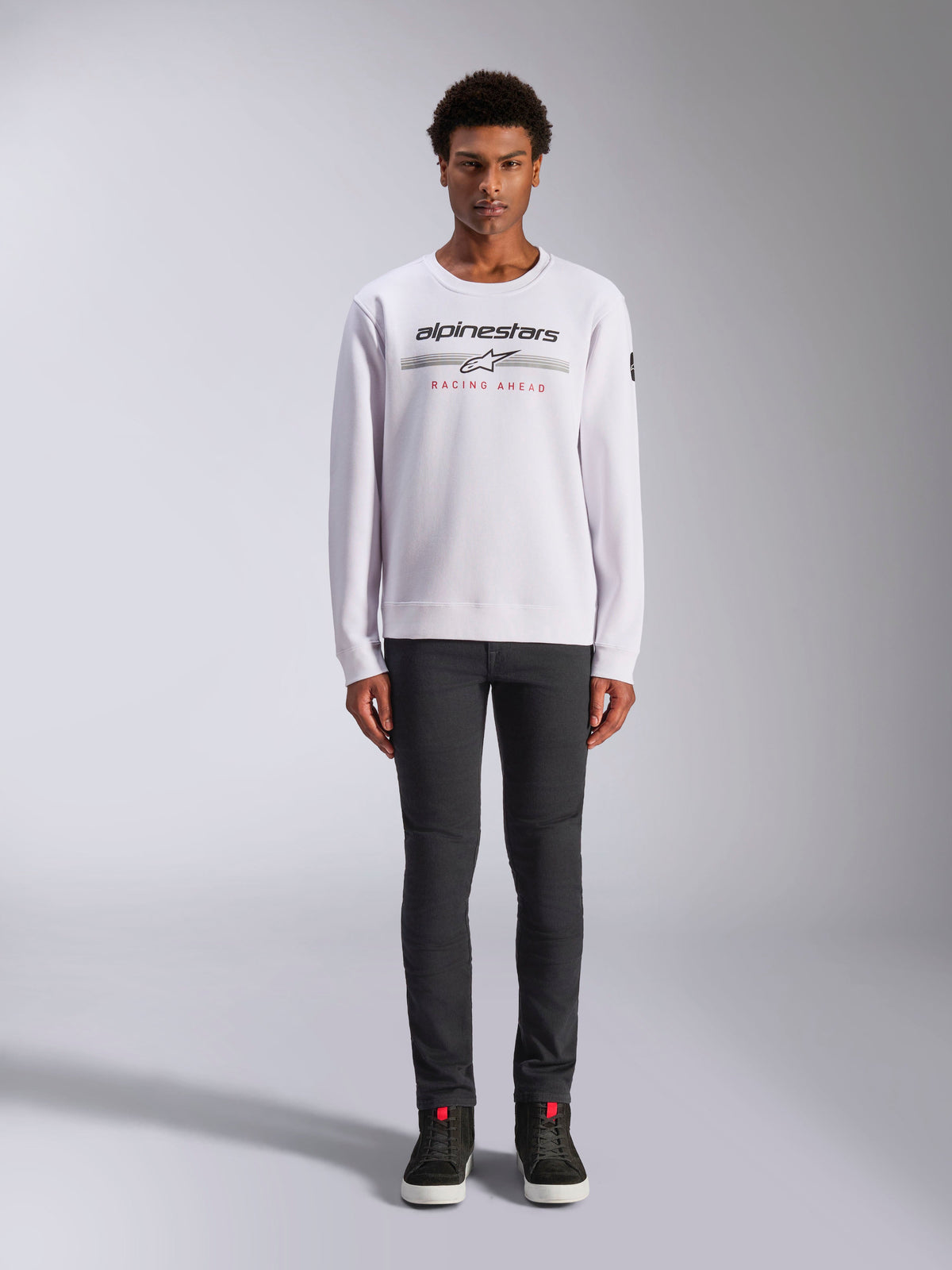 Alpinestars Bettern Crew, sweat à col rond blanc, porté par un mannequin homme avec un pantalon gris foncé et des baskets montantes noires, avec le logo Alpinestars et le texte rouge 'Racing Ahead', style streetwear décontracté inspiré du sport automobile