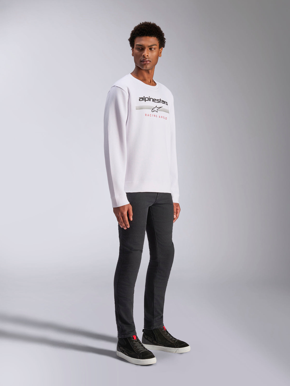 Alpinestars Bettern Crew, sweat-shirt blanc à col rond avec logo Alpinestars noir et texte rouge 'Racing Ahead', style streetwear décontracté inspiré du sport automobile, porté par un mannequin avec un pantalon sombre et des baskets noires
