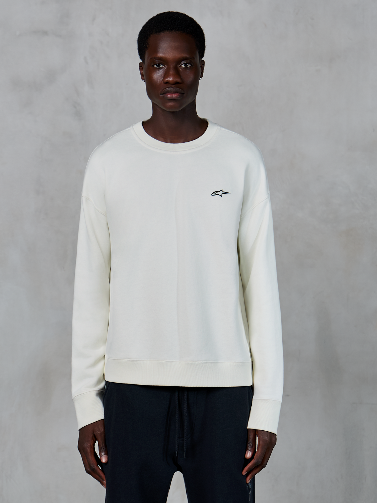 Alpinestars Essentials Ageless Crew, Off White sweatshirt met ronde hals, klein zwart Astars-logo op de linkerborst, casual lifestyle sportkleding, premium fleece textuur, gedragen door model met zwarte joggingbroek