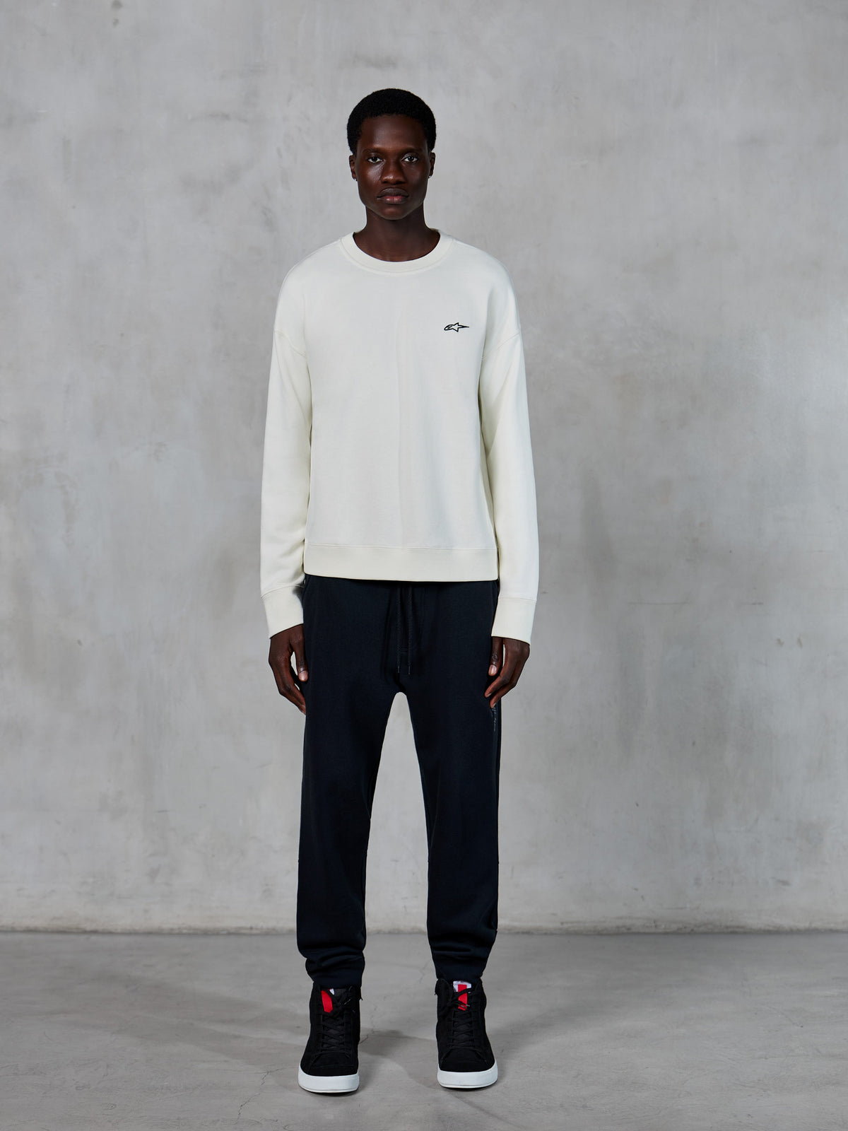 Alpinestars Essentials Ageless Crew, crewneck sweatshirt, Off White met klein zwart sterlogo, gedragen door model met zwarte joggers, casual op motorsport geïnspireerde streetwear