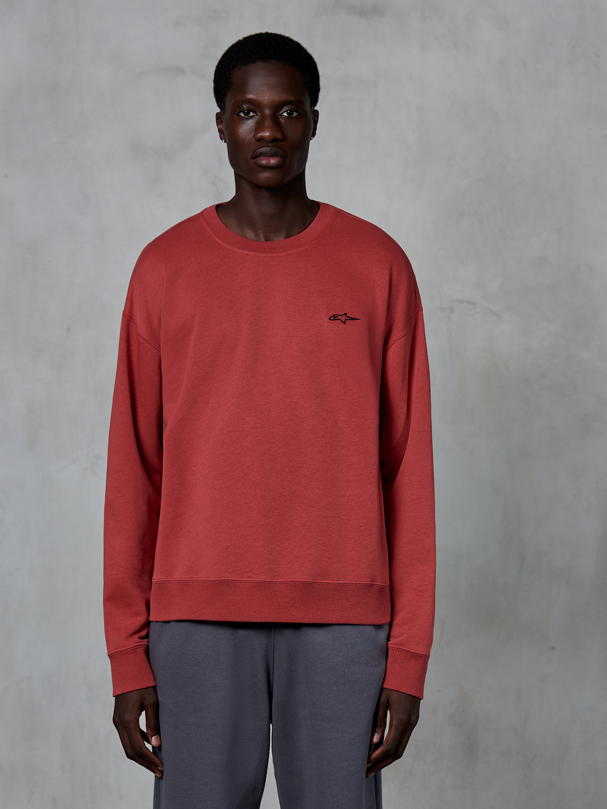 Alpinestars Essentials Ageless Crew, sweat-shirt à col rond, rouge brique, petit logo noir sur la poitrine, porté par un mannequin avec un pantalon gris, polaire douce de qualité supérieure, streetwear décontracté
