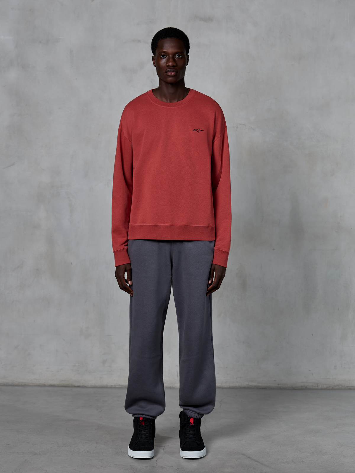 Alpinestars Essentials Ageless Crew, sweat-shirt à col rond rouge brique, porté avec un pantalon de jogging gris et des baskets noires, avec un petit logo noir sur la poitrine, streetwear décontracté d'inspiration sport mécanique