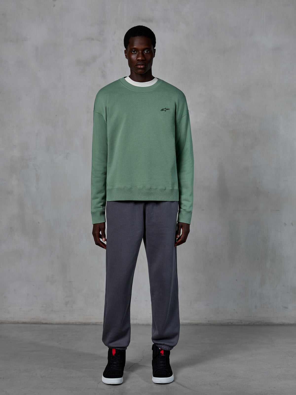 Alpinestars Essentials Ageless Crew, sweat-shirt à col rond en vert Cactus, doté d'un petit logo noir sur la poitrine, porté par un mannequin avec un pantalon de jogging gris et des baskets noires, streetwear décontracté inspiré du sport automobile